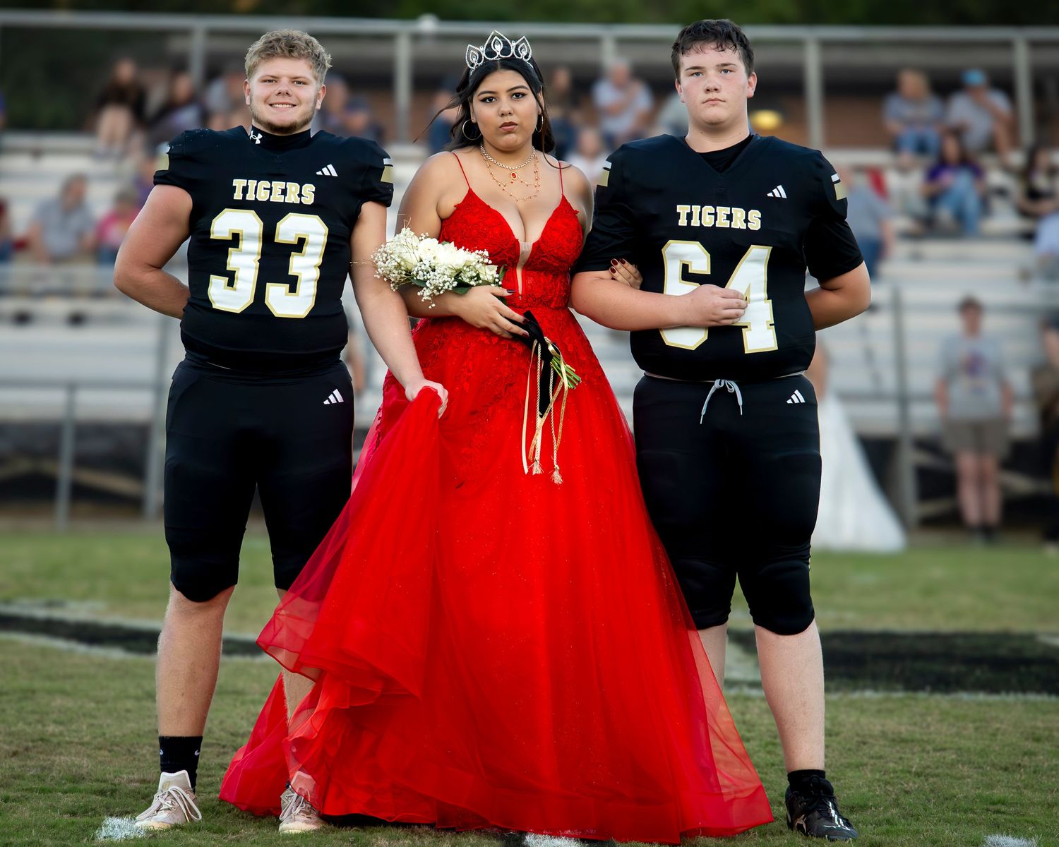 A person in a flowing red formal dress stands on a football field among players in black uniforms during a special event.
