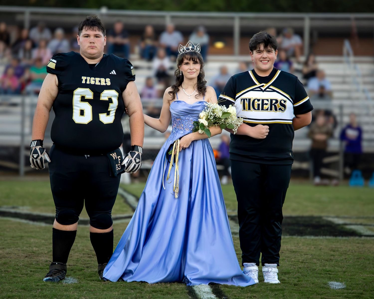 Football players in black uniforms pose with someone wearing a light blue formal dress on a sports field.