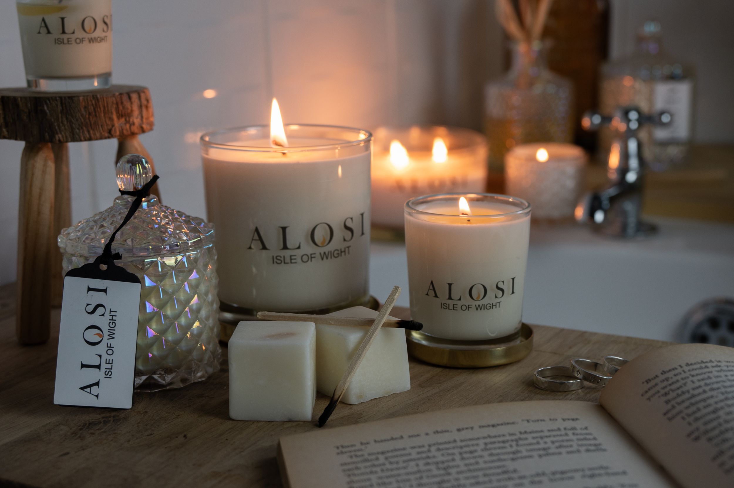 Glowing candles in glass jars with ALOS branding create a cozy atmosphere on a rustic wooden surface.