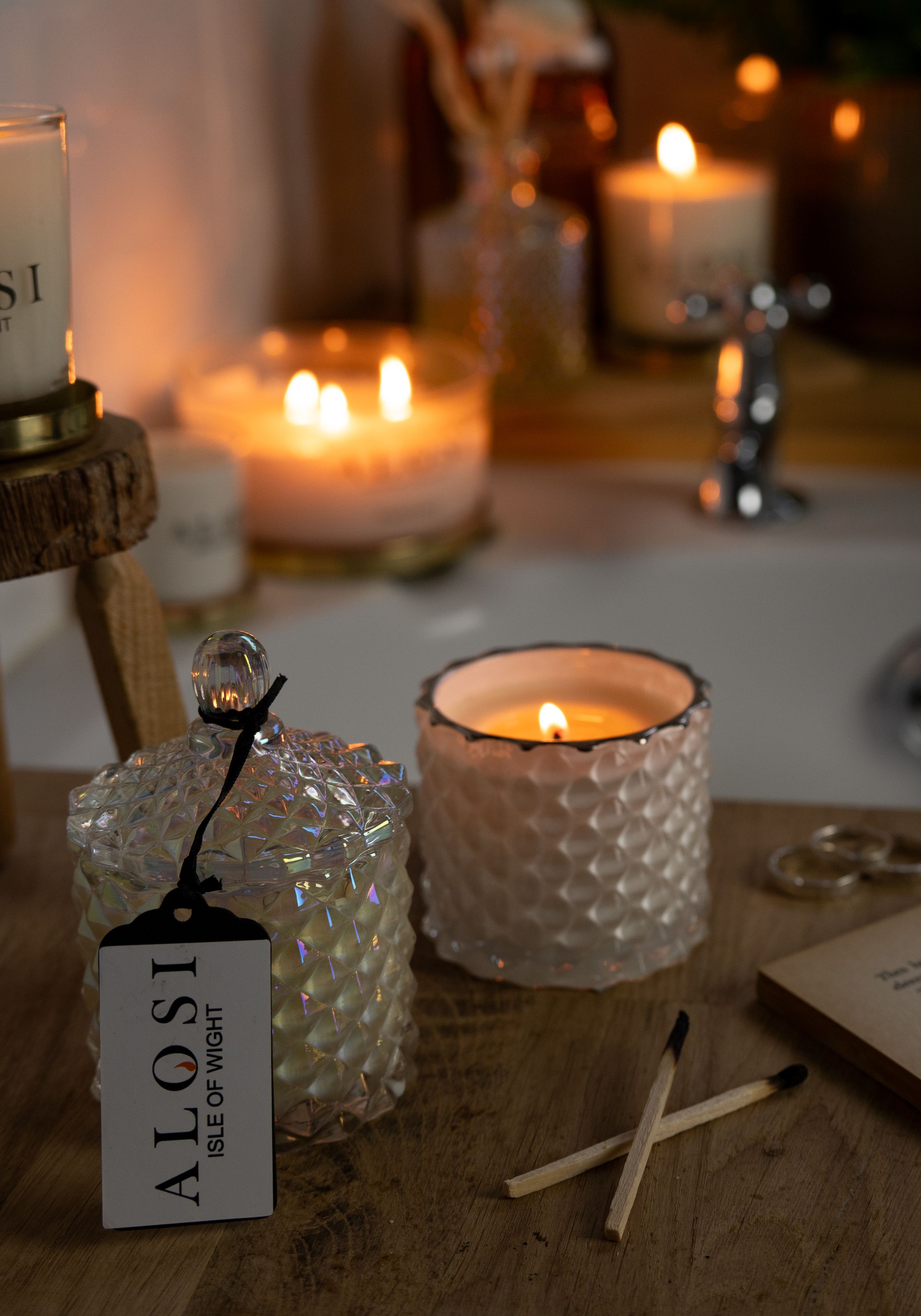 Lit Alosi candles in textured glass holders with hanging tags create an intimate ambiance on a rustic wooden surface.
