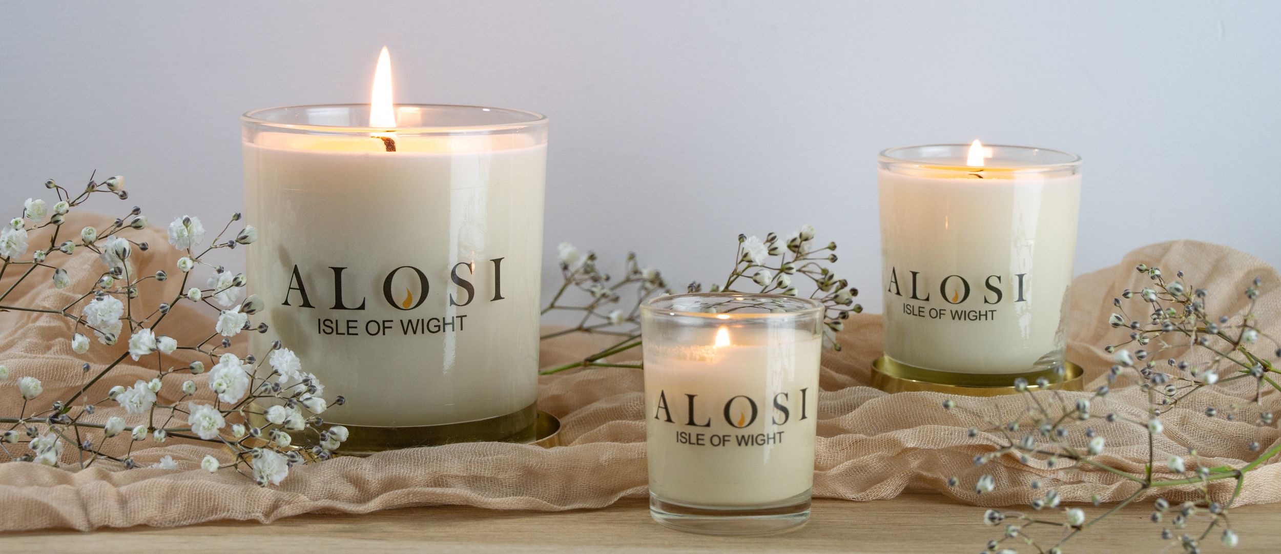 Elegant white candle in glass jar with delicate floral decorations and soft glowing flame against light background.