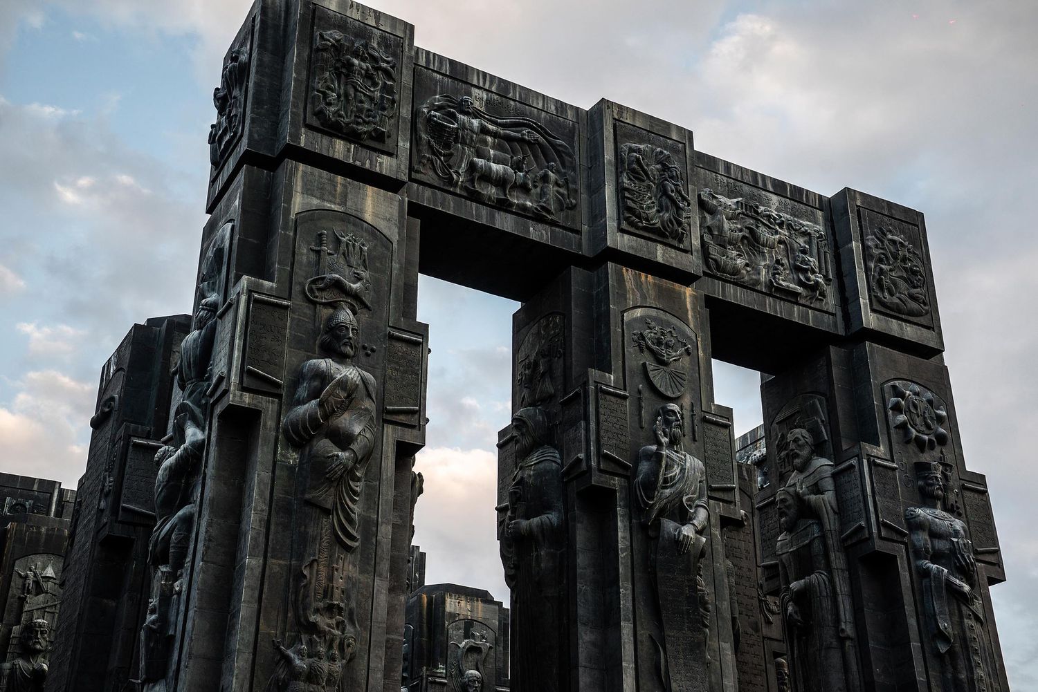 Ancient stone carvings on a monumental wall show intricate Mayan hieroglyphic designs against a cloudy sky.
