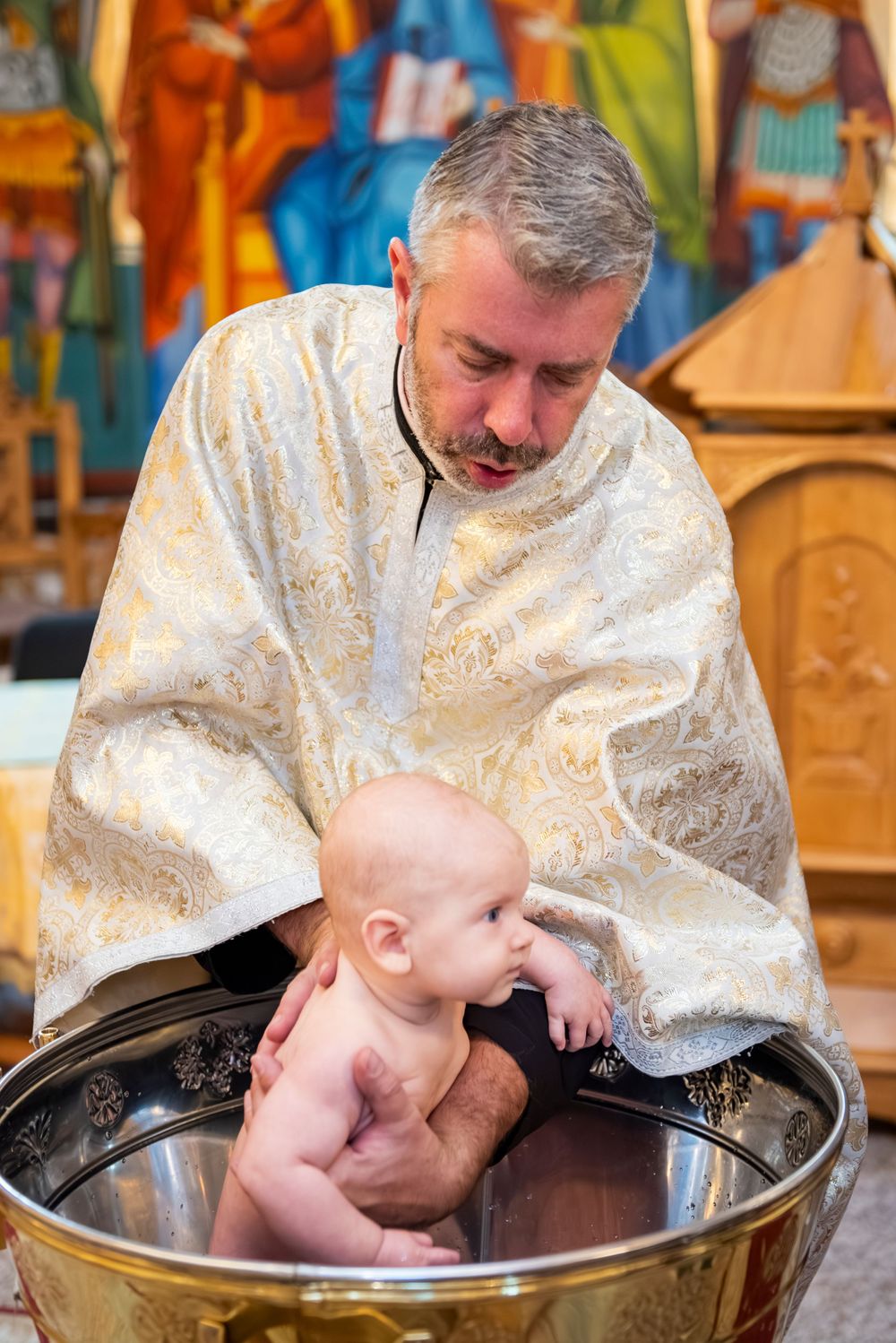 Orthodox Christian baptism ceremony showing priest performing infant baptism in traditional font.