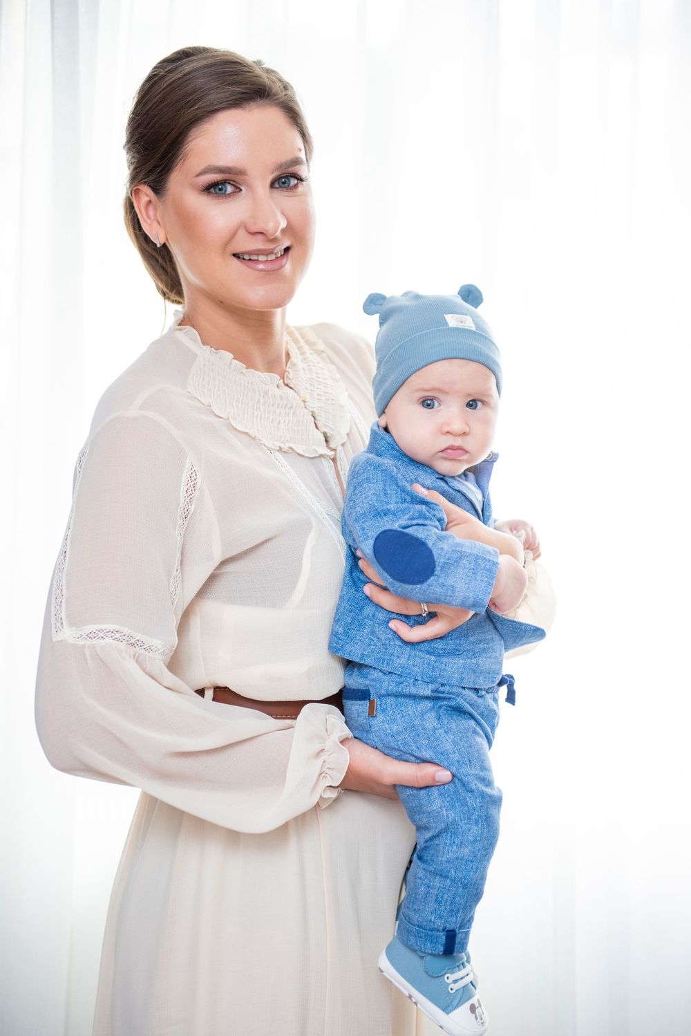 A mother and baby wearing coordinated white and blue outfits pose together in a bright indoor setting.