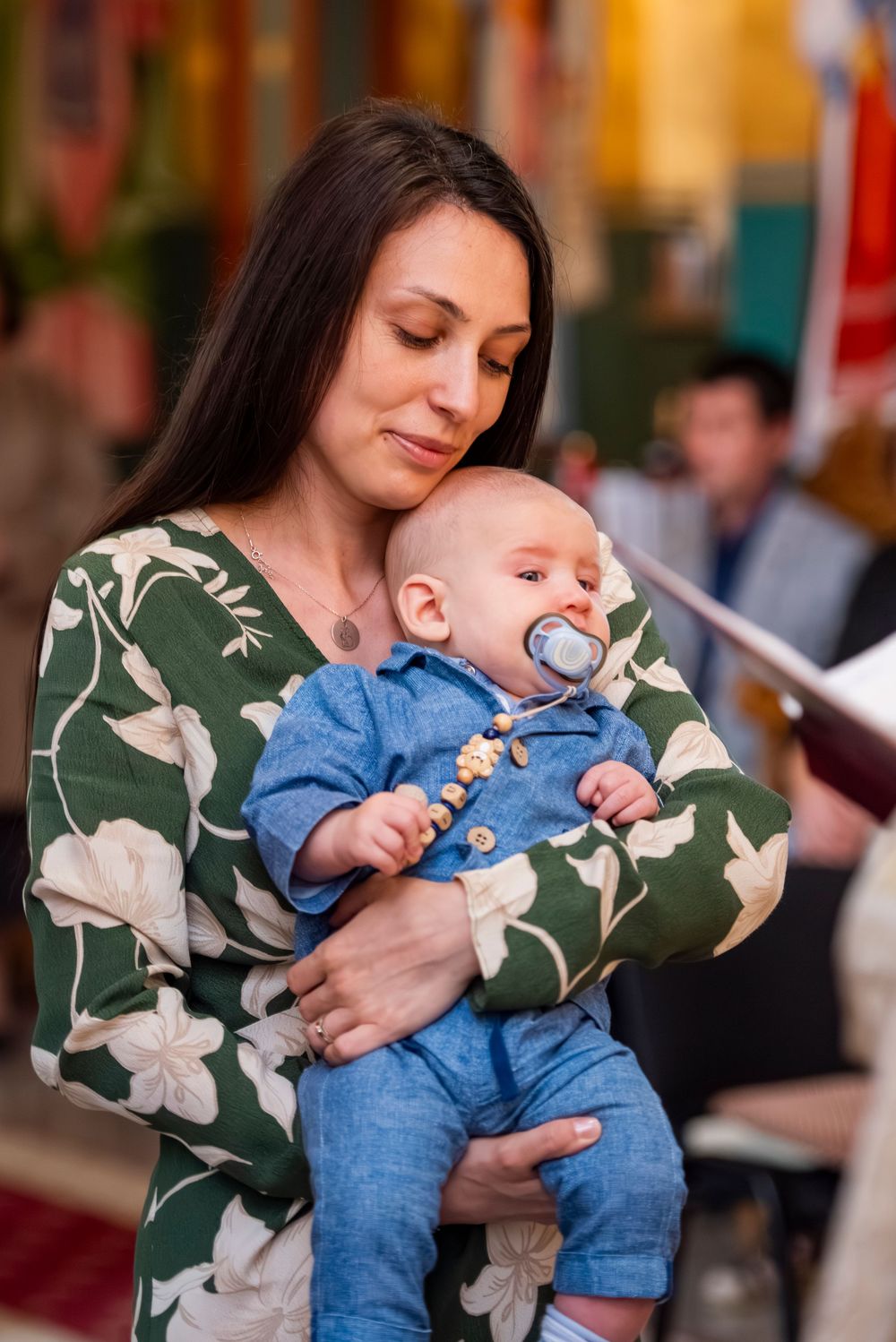 Mother in floral green dress holds and comforts infant wearing blue in outdoor setting.
