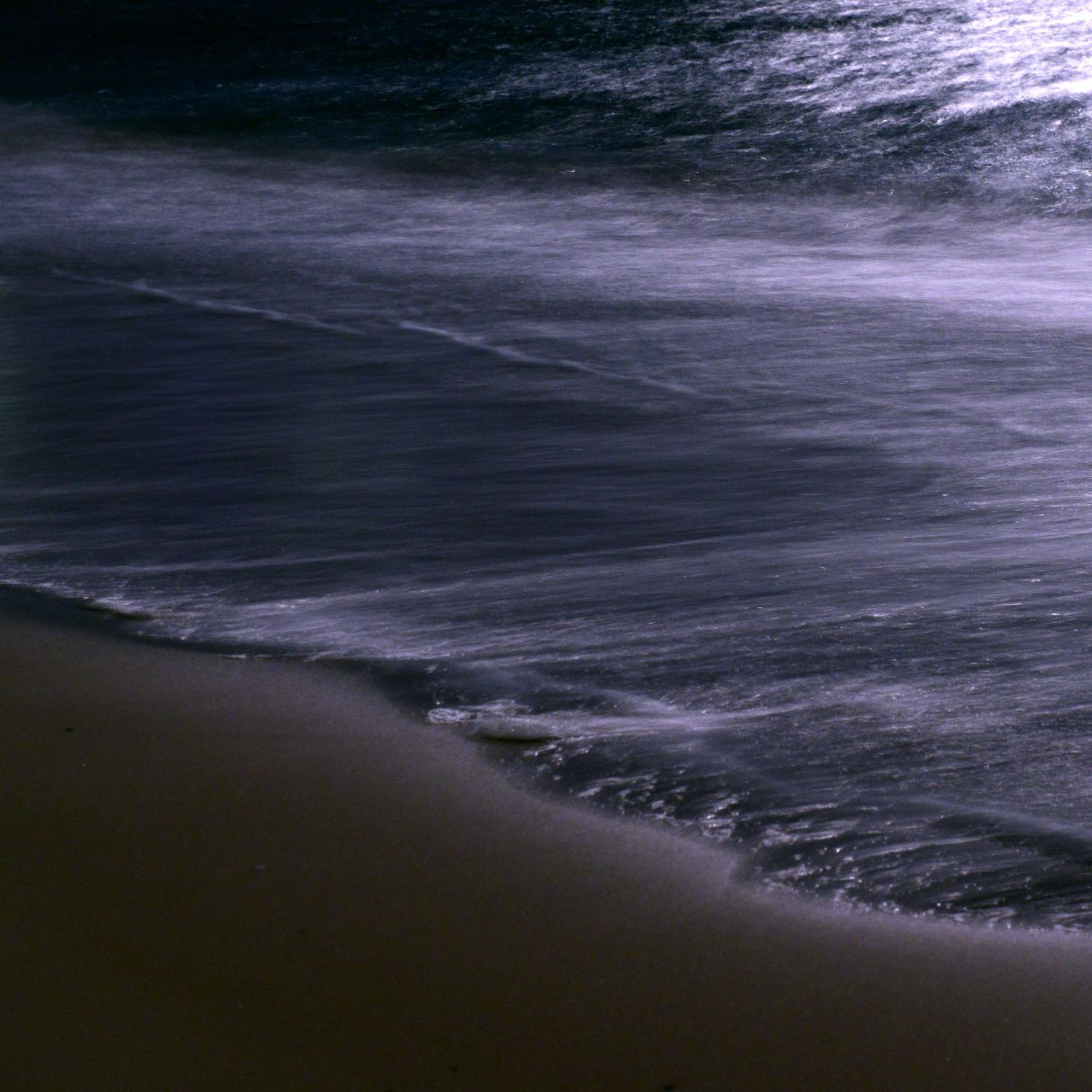 Dark waves roll onto a sandy beach at night with a moody, dramatic sky overhead.