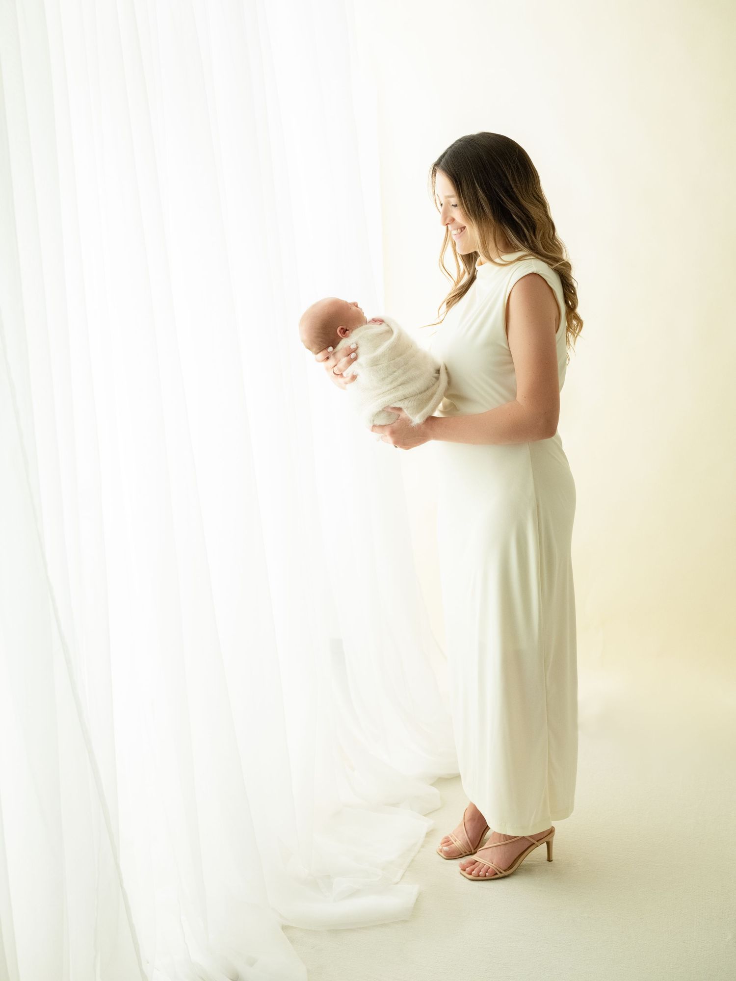 A serene portrait showing a parent in a white dress cradling a swaddled newborn baby by a sunlit window.