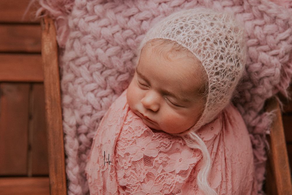Newborn baby wearing delicate lace bonnet rests peacefully on pink blanket in rustic wooden setting.