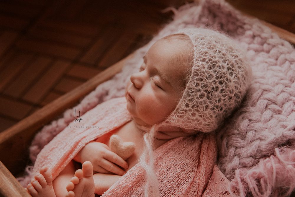 A baby wearing a white lace bonnet rests peacefully on pink bedding in warm lighting.