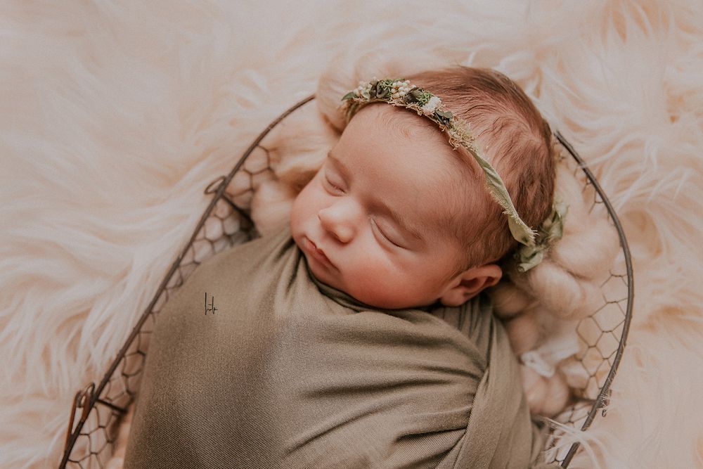 A sleeping newborn baby wearing a delicate floral crown rests peacefully on soft beige fabric.