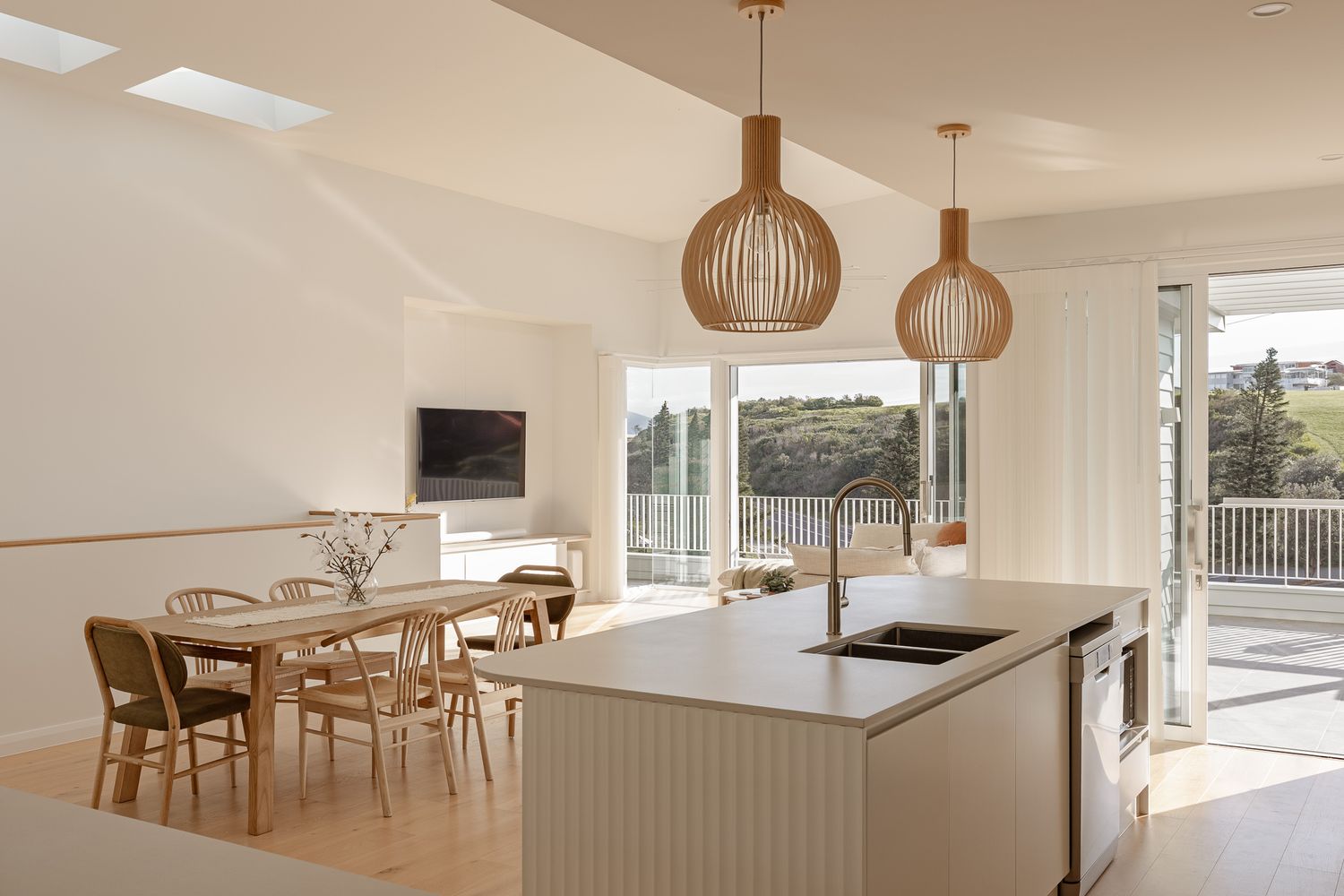 Modern open concept kitchen and dining area with wooden pendant lights and white cabinetry overlooking scenic views.