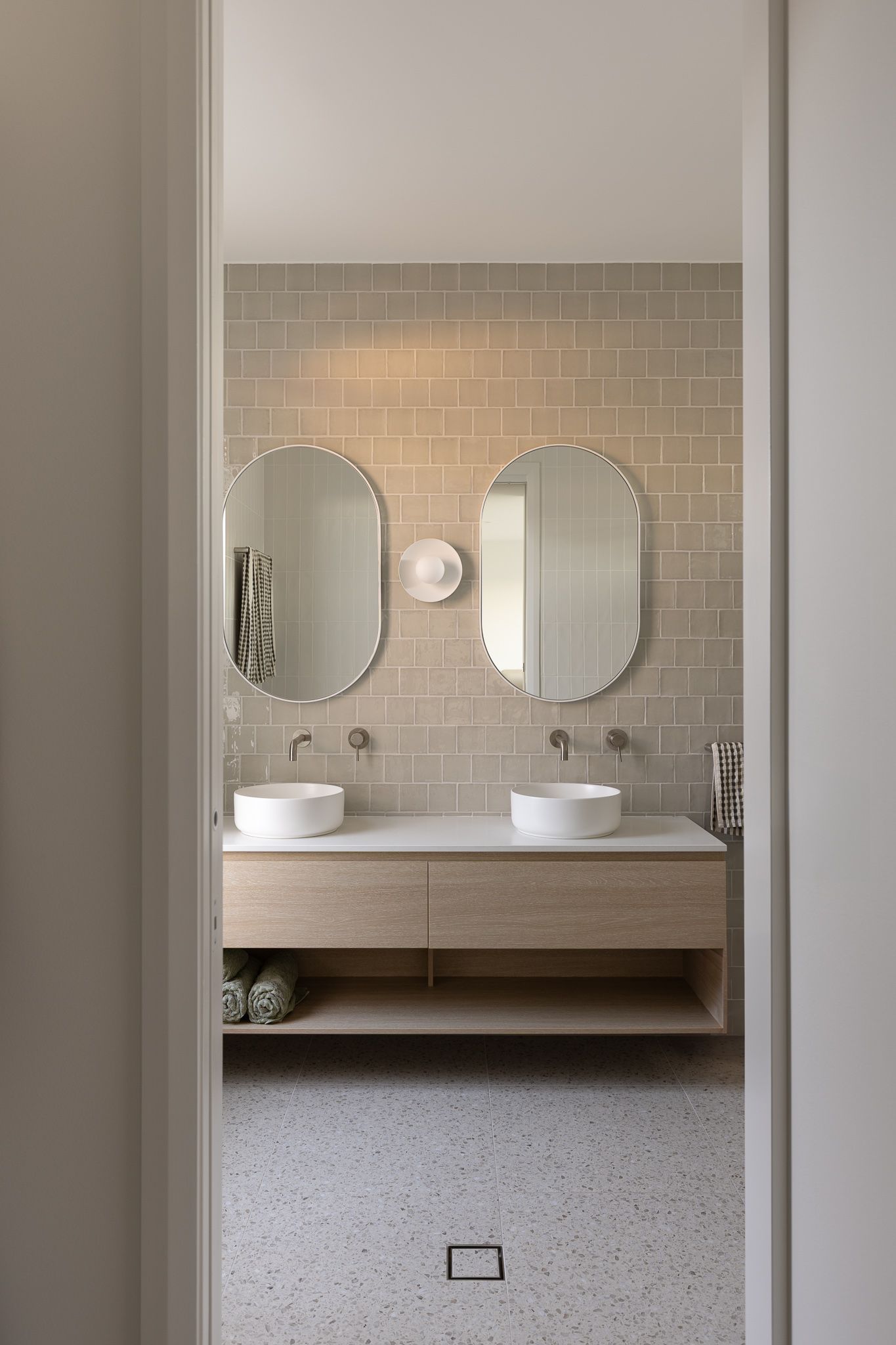 Modern bathroom interior with dual round mirrors, floating wooden vanity, and white vessel sinks against beige tile wall.