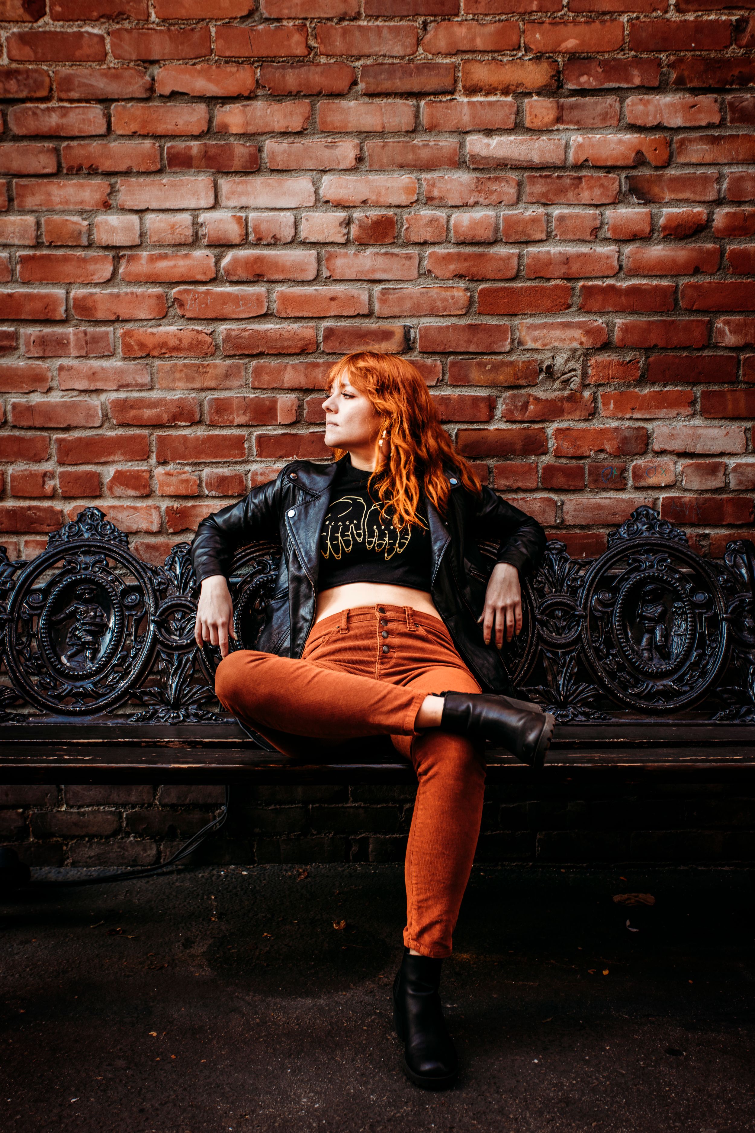 A redhead woman in a leather jacket and rust orange pants sits on an ornate bench against a brick wall.