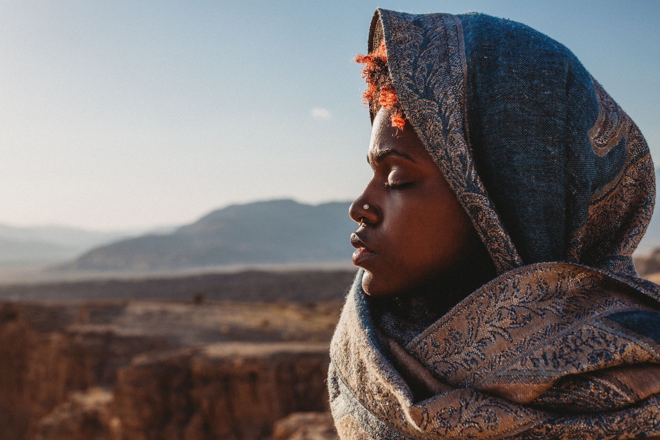 A young Black woman wearing a decorative hijab stands against a desert mountain backdrop in golden light.