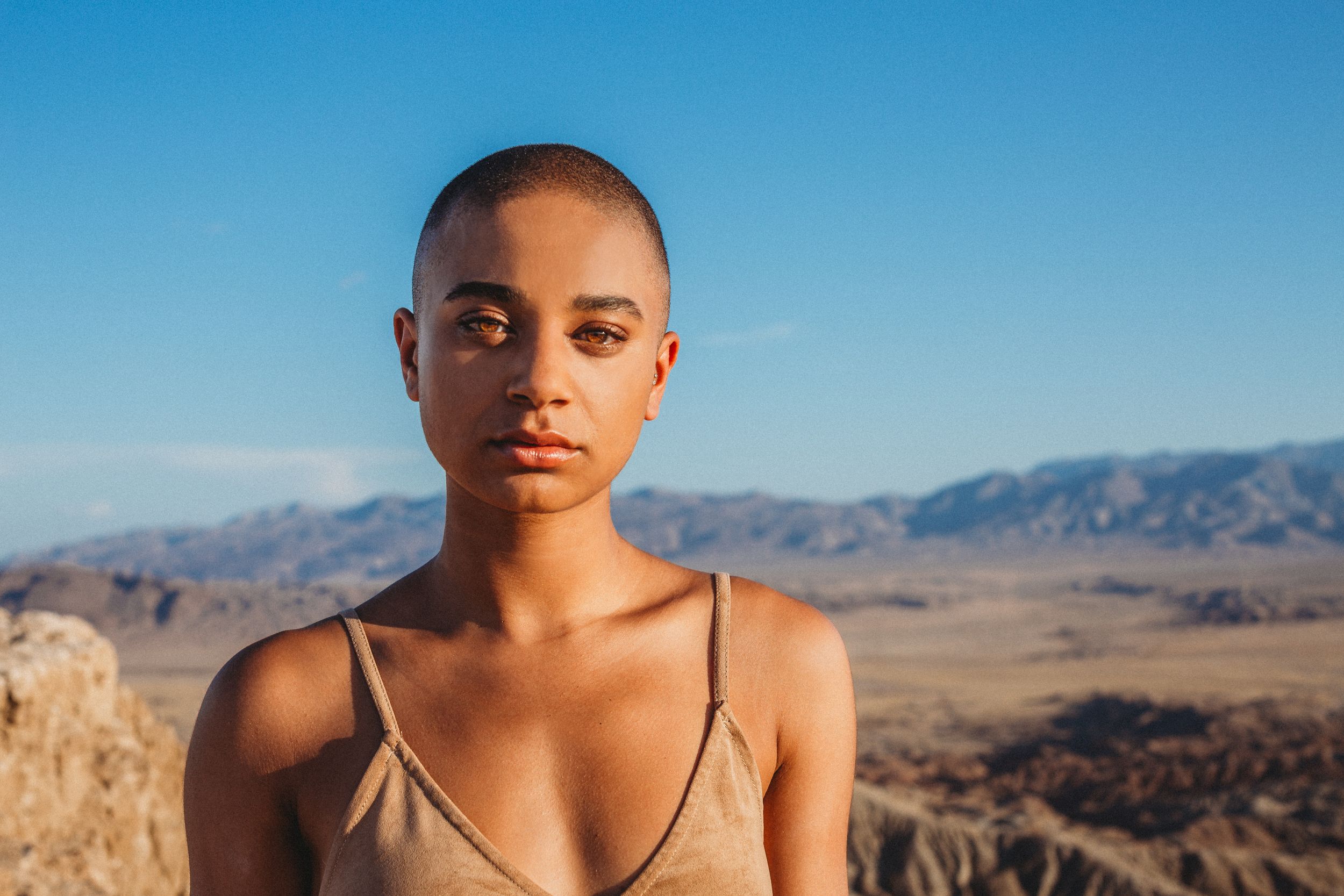 A bald woman in a tan top poses confidently against a vast desert mountain backdrop under blue sky.