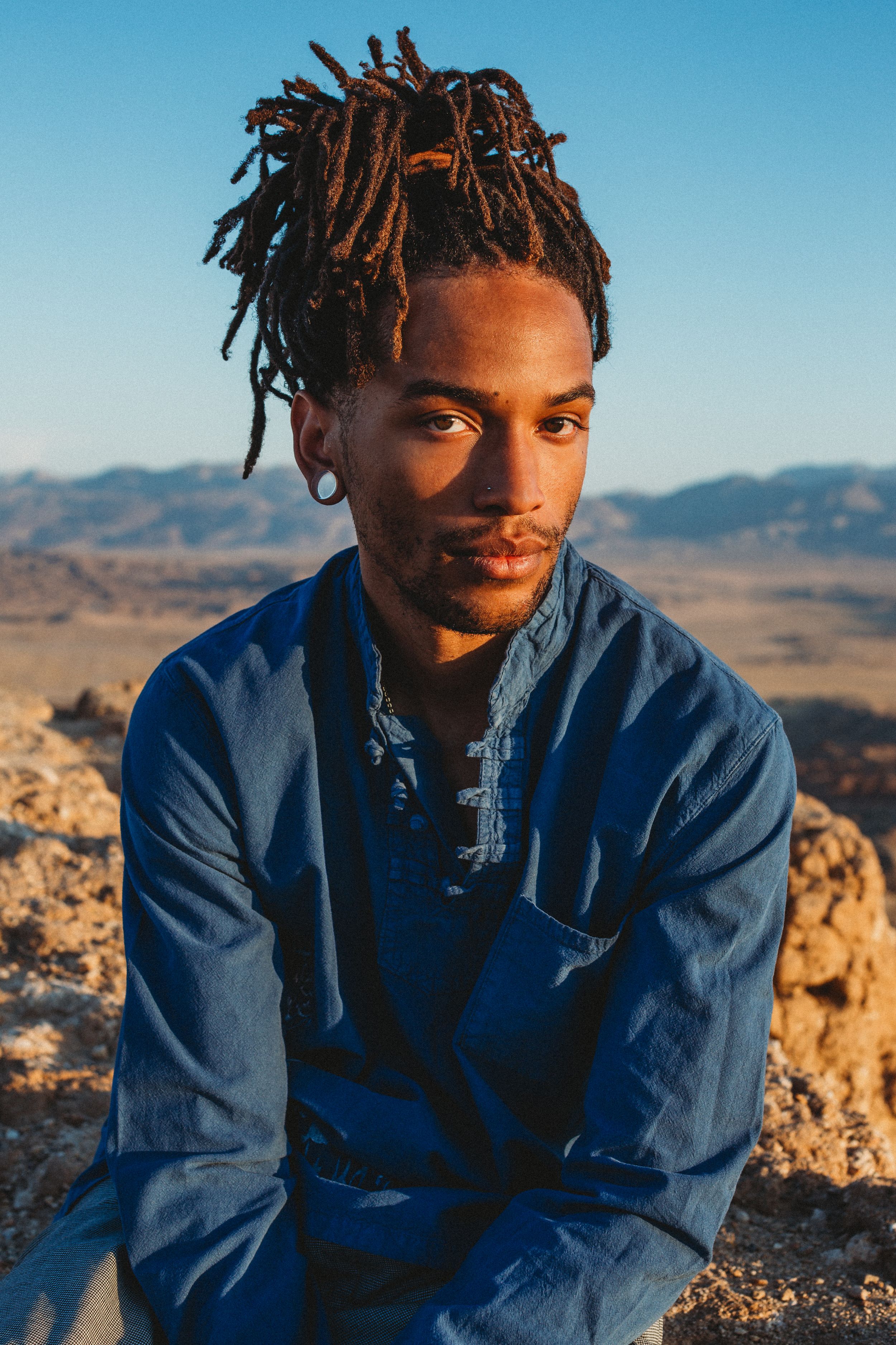 A young Black man with dreadlocks wearing a blue denim shirt sits on rocks with a desert landscape behind him.