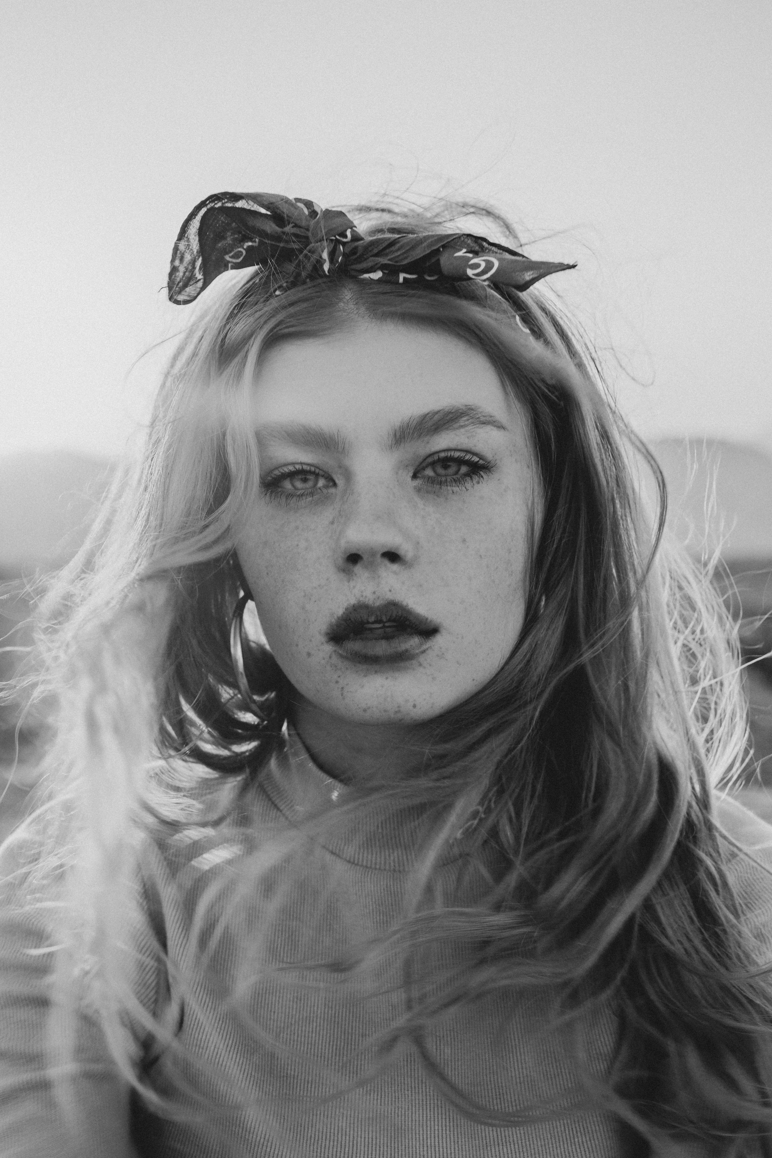 A young woman with freckles and a bandana headband gazes forward in a black and white outdoor portrait.