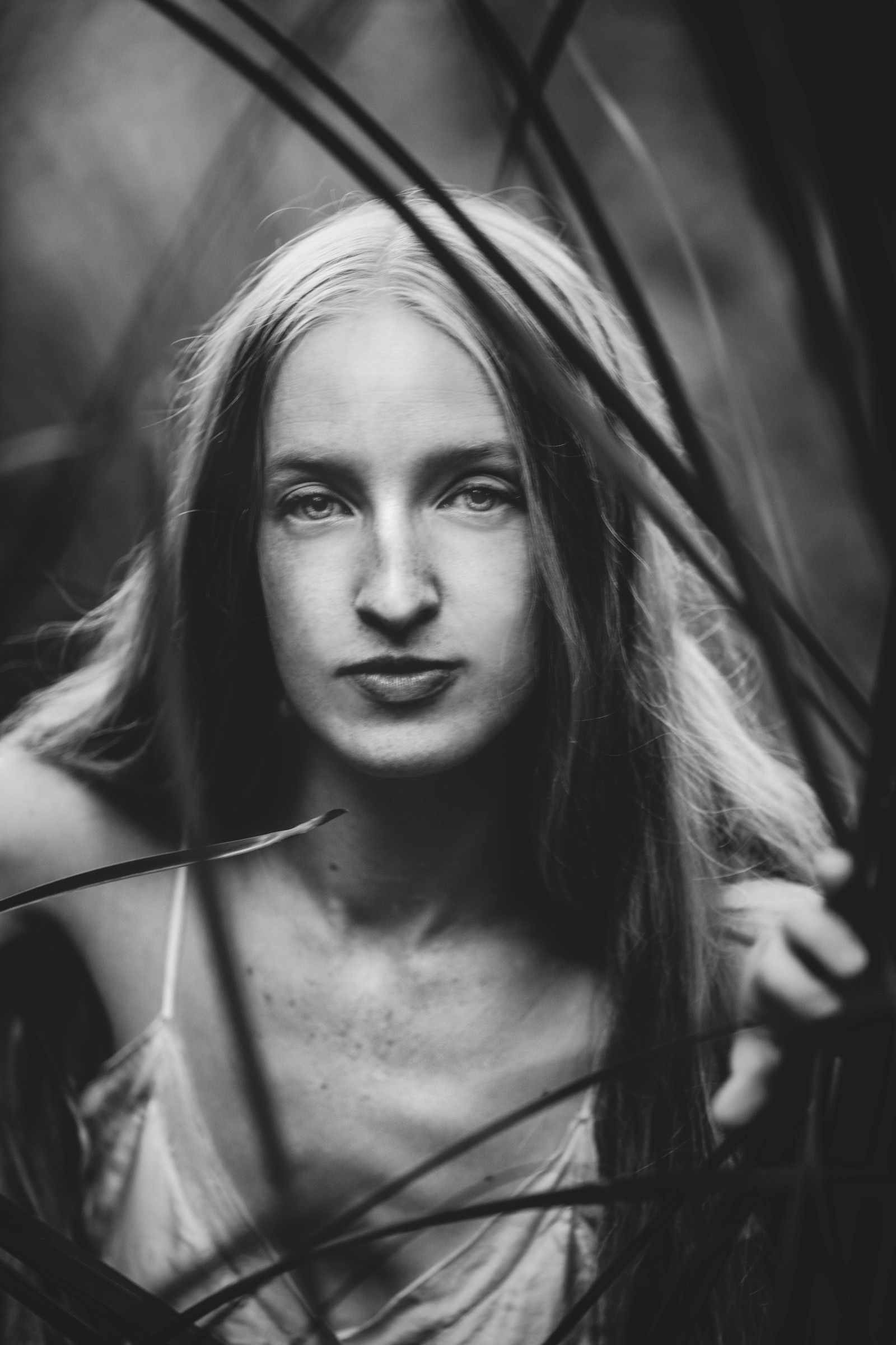 A black and white portrait of a blonde woman peering through tall grass blades in a natural setting.