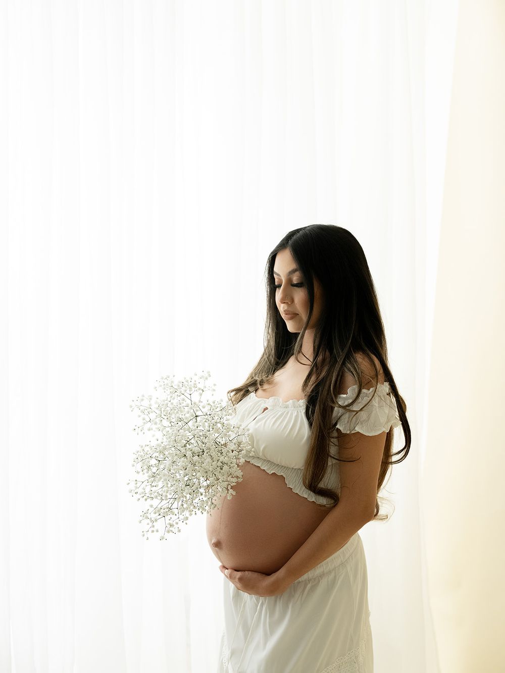 Maternity photo series showing a woman in white dress holding baby's breath flowers against a light background.