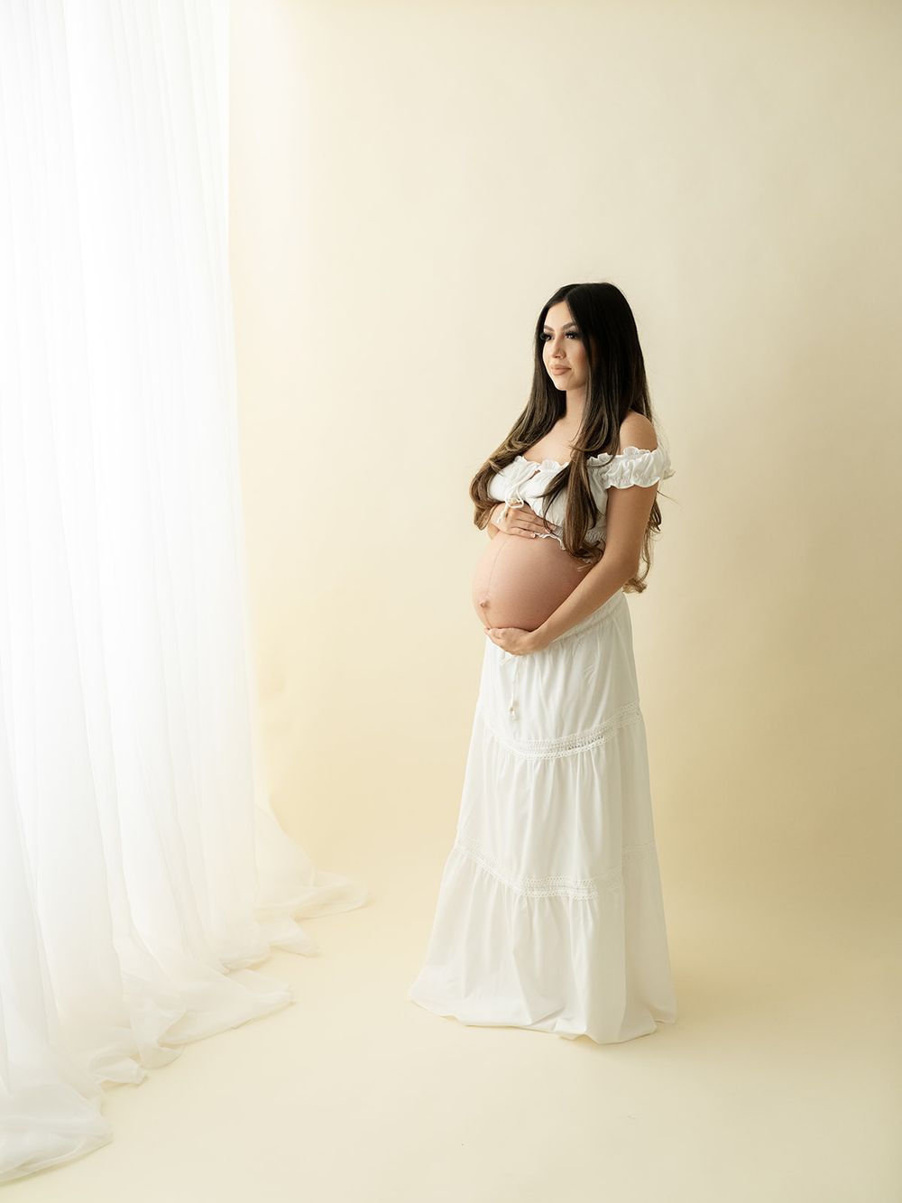 A maternity photo series showing a pregnant woman in a flowing white dress against a light beige backdrop.