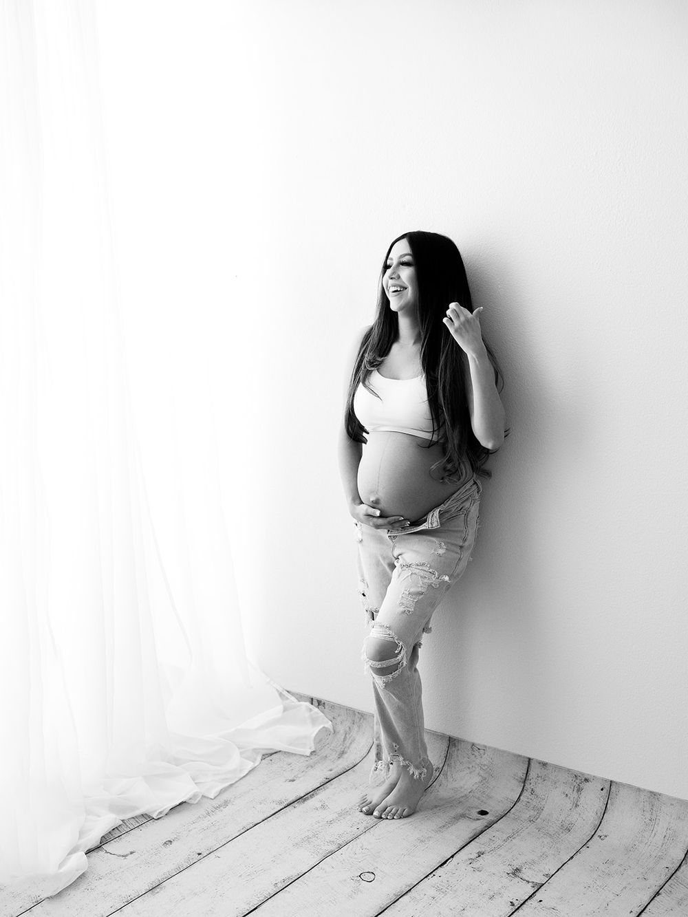 Black and white candid maternity photo showing pregnant woman in casual attire against white wall.