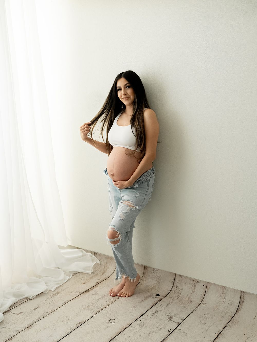 A maternity photoshoot featuring a white crop top and distressed jeans against a rustic white wooden floor backdrop.