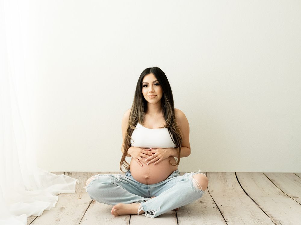 Expectant mother in casual jeans and white top sitting on rustic wooden floor during maternity photoshoot.