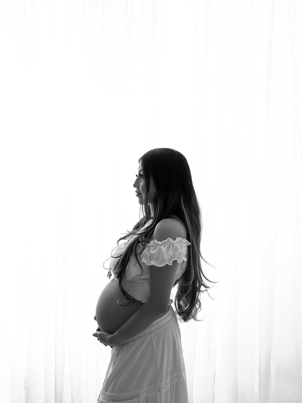 A silhouette of a woman in a white dress stands near a window in a black and white artistic portrait.