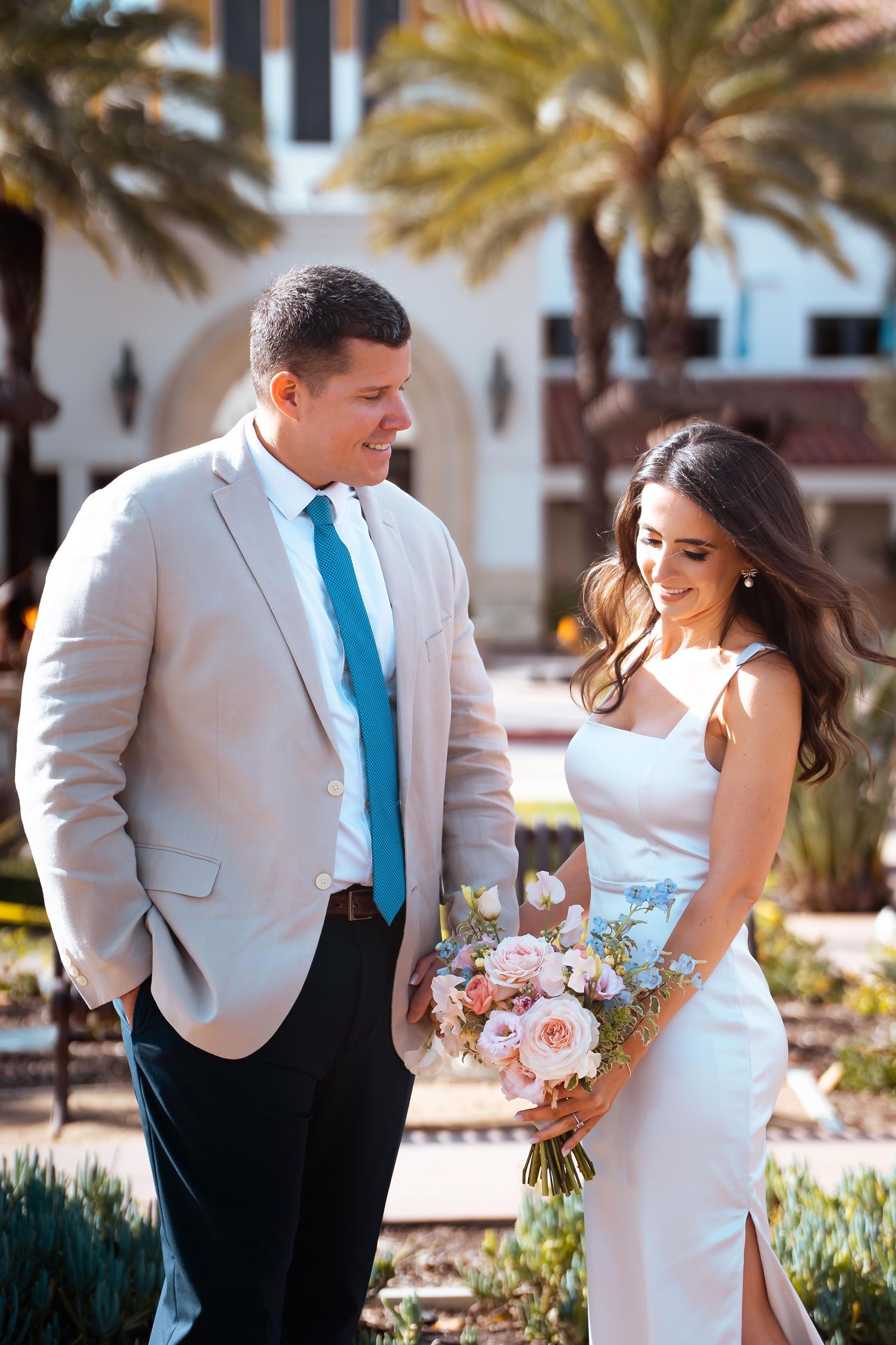 A couple in formal attire shares a joyful moment in a tropical setting with palm trees and a pink floral bouquet.