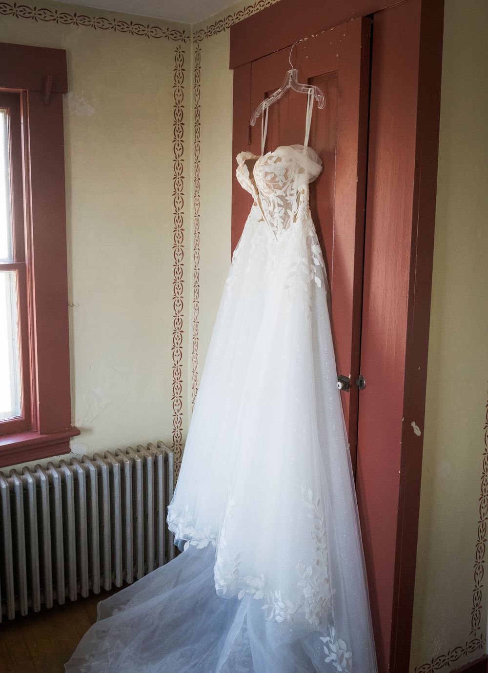 White lace wedding dress hanging on display against a red wall with vintage radiator.