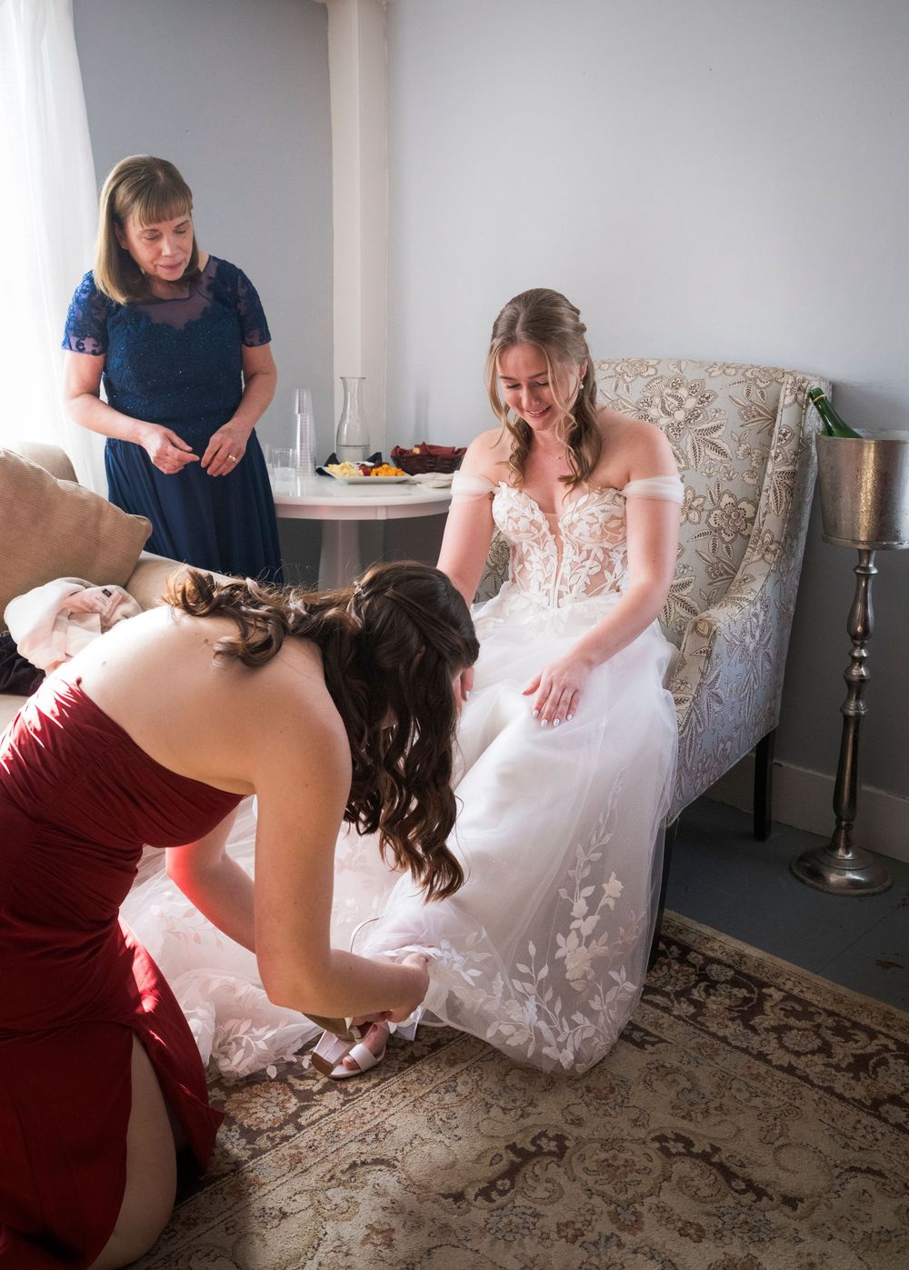 A bride in a white lace dress sits in an elegant chair while being helped with her shoes before the wedding ceremony.