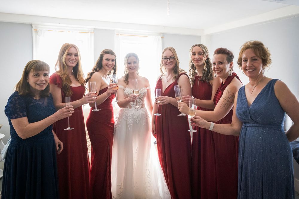 A bridal party in burgundy and navy dresses toasts with champagne glasses in a bright room.