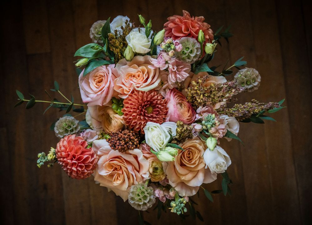 A romantic bouquet of pink and coral roses with dahlias and greenery arranged on dark wood background.