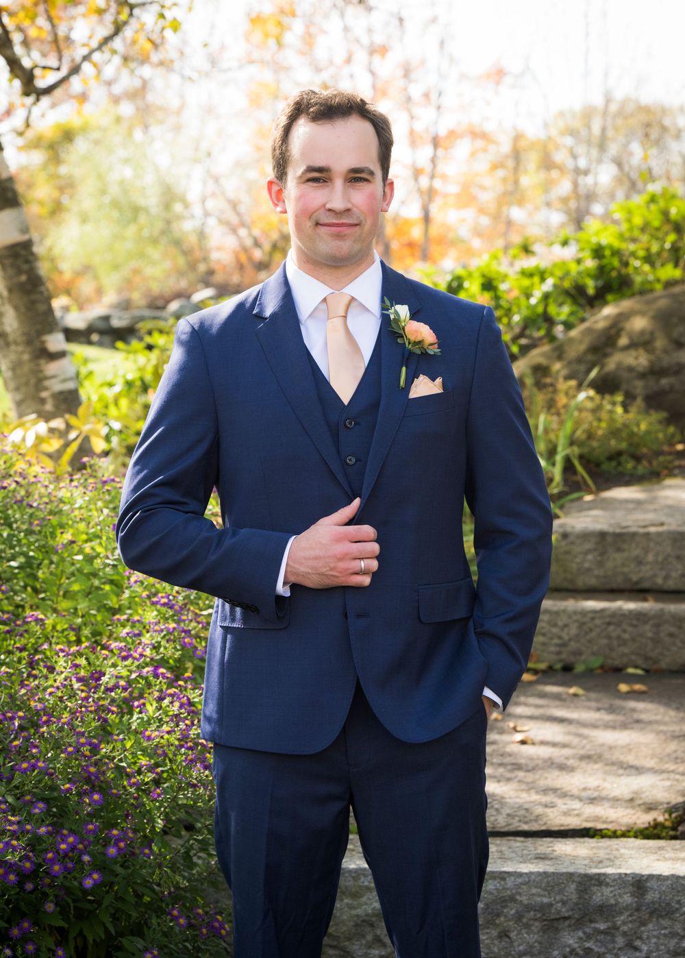 A formal navy blue three-piece suit with peach tie and boutonniere in an autumn garden setting.
