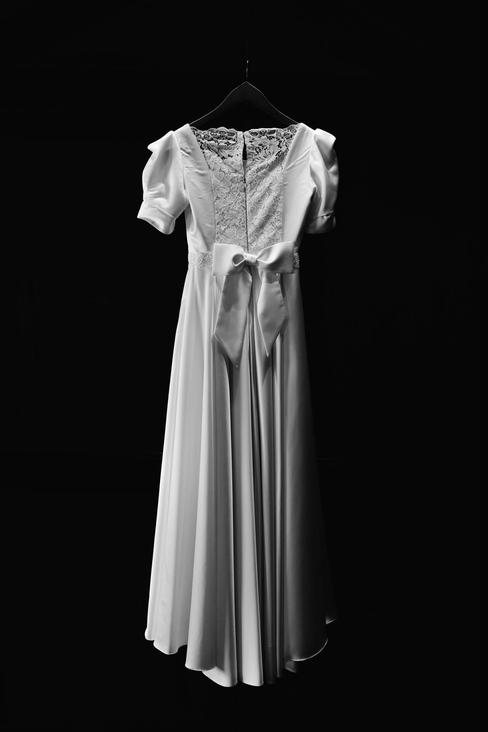 Elegant white vintage-style wedding dress with lace details and short puffed sleeves hanging against black background.