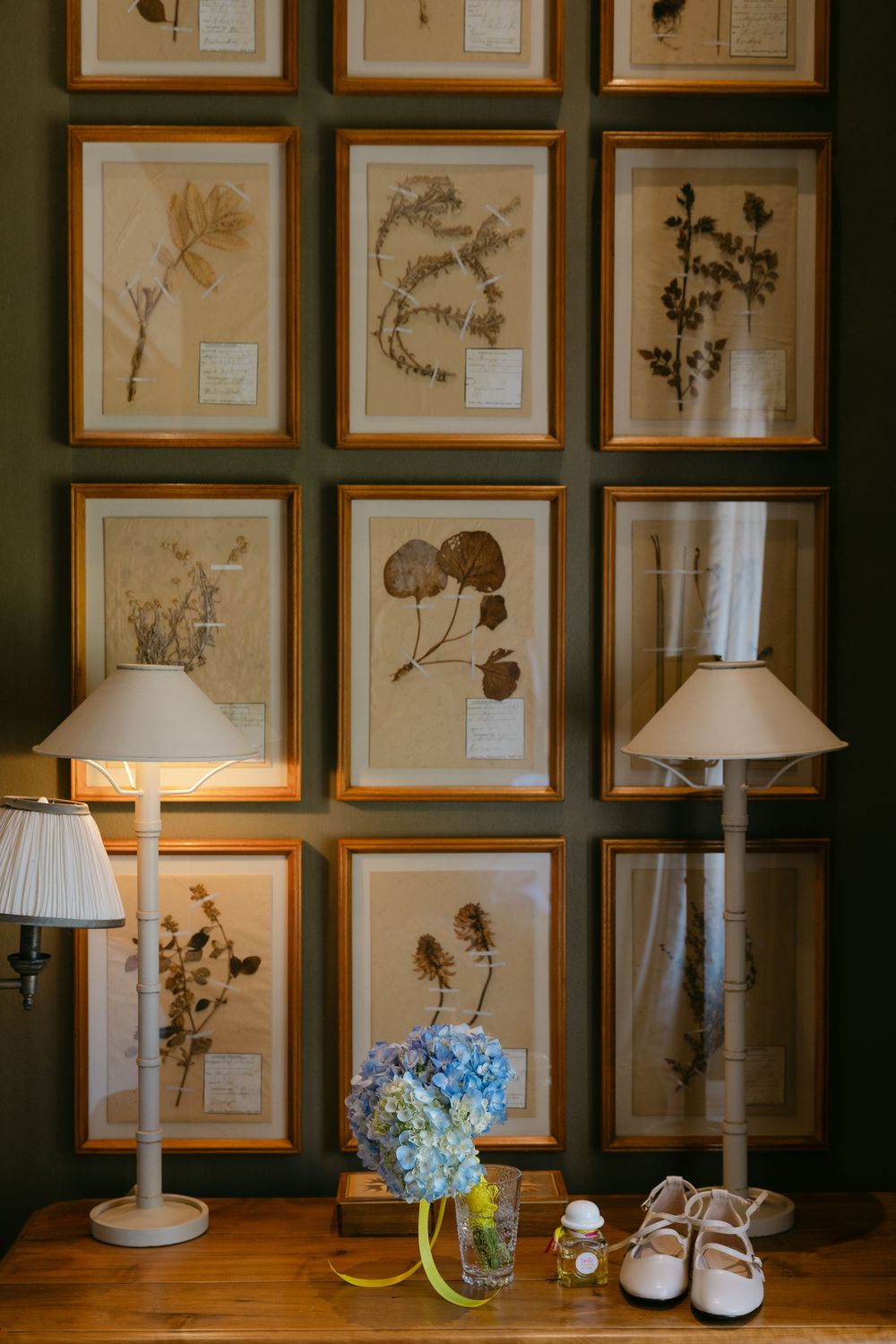 Vintage botanical prints in wooden frames line a wall above a table with white lamps and blue hydrangeas.