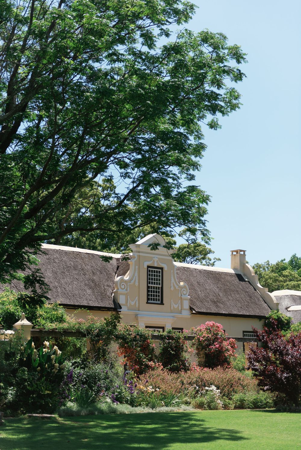 Rustic thatched-roof cottage with blooming gardens and trees against a blue summer sky.