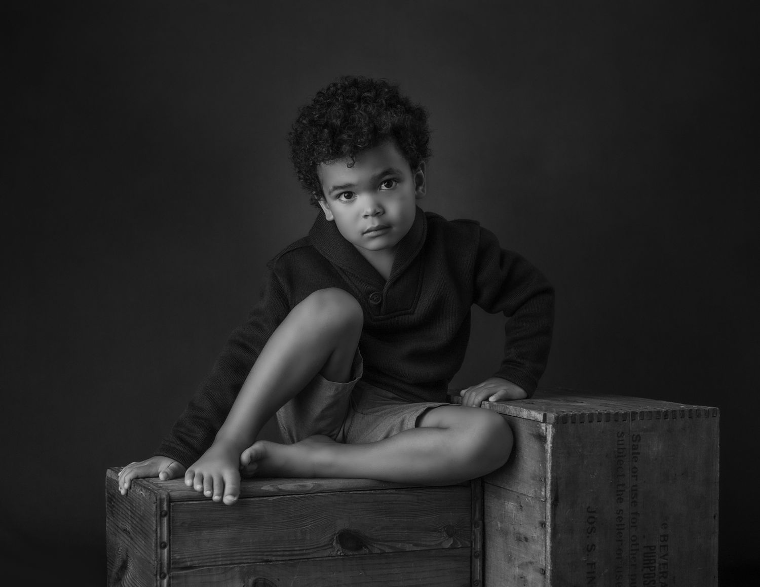 Black and white portrait of a young boy sitting cross-legged on a wooden crate.