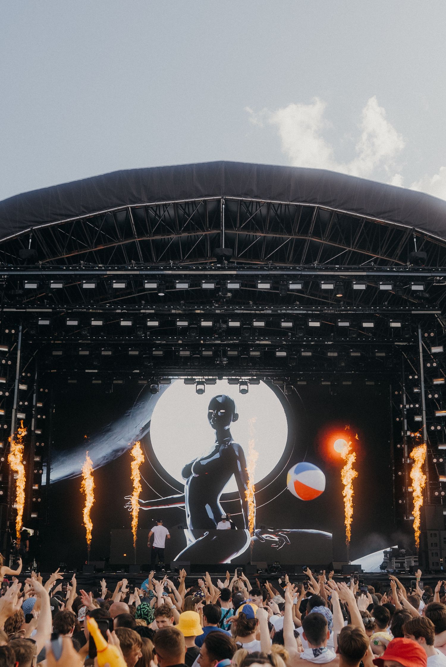 Concert stage with pyrotechnics and large video screen displaying silhouette performer to cheering crowd.