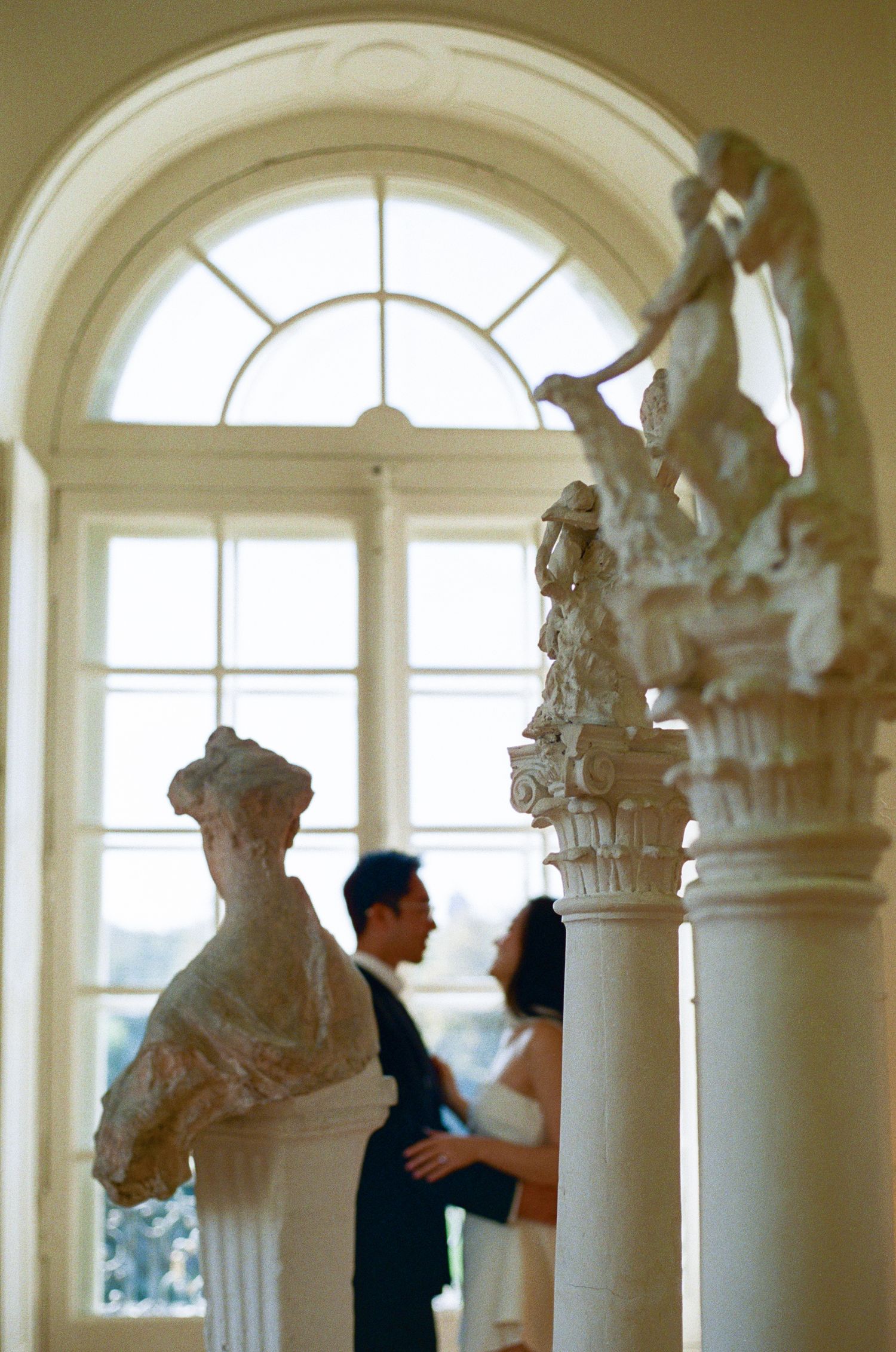 A couple shares an intimate moment in an elegant room with arched windows and classical architectural details.