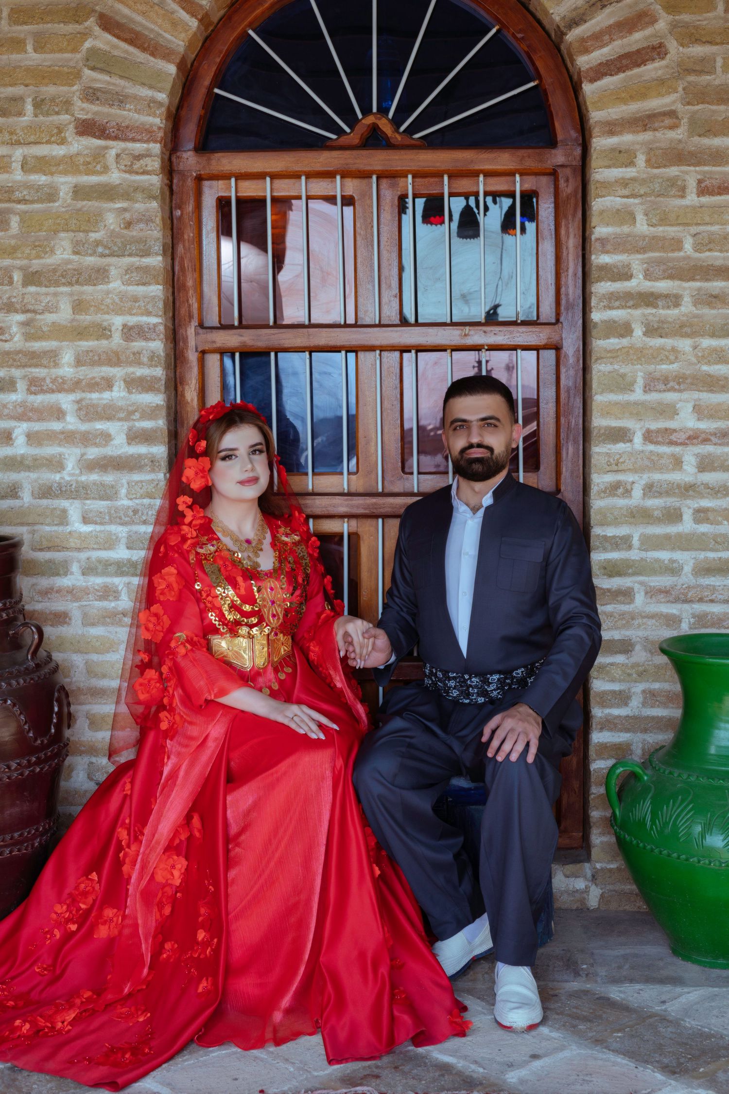 A couple in traditional attire poses on a brick porch, with the woman wearing a vibrant red dress and the man in a dark suit.