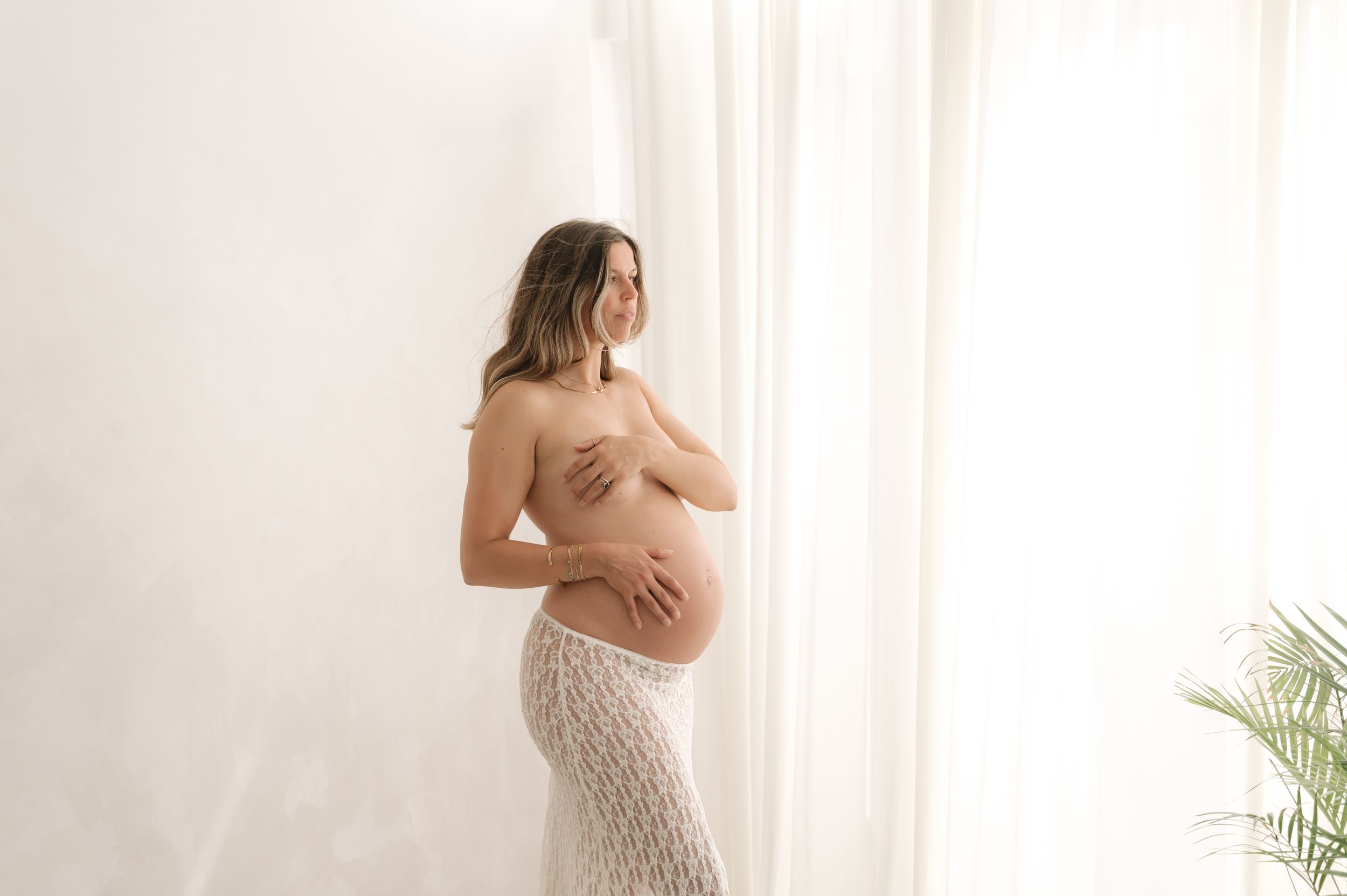Pregnant woman in lace skirt posing topless against white wall near sheer curtains in a maternity photo shoot.