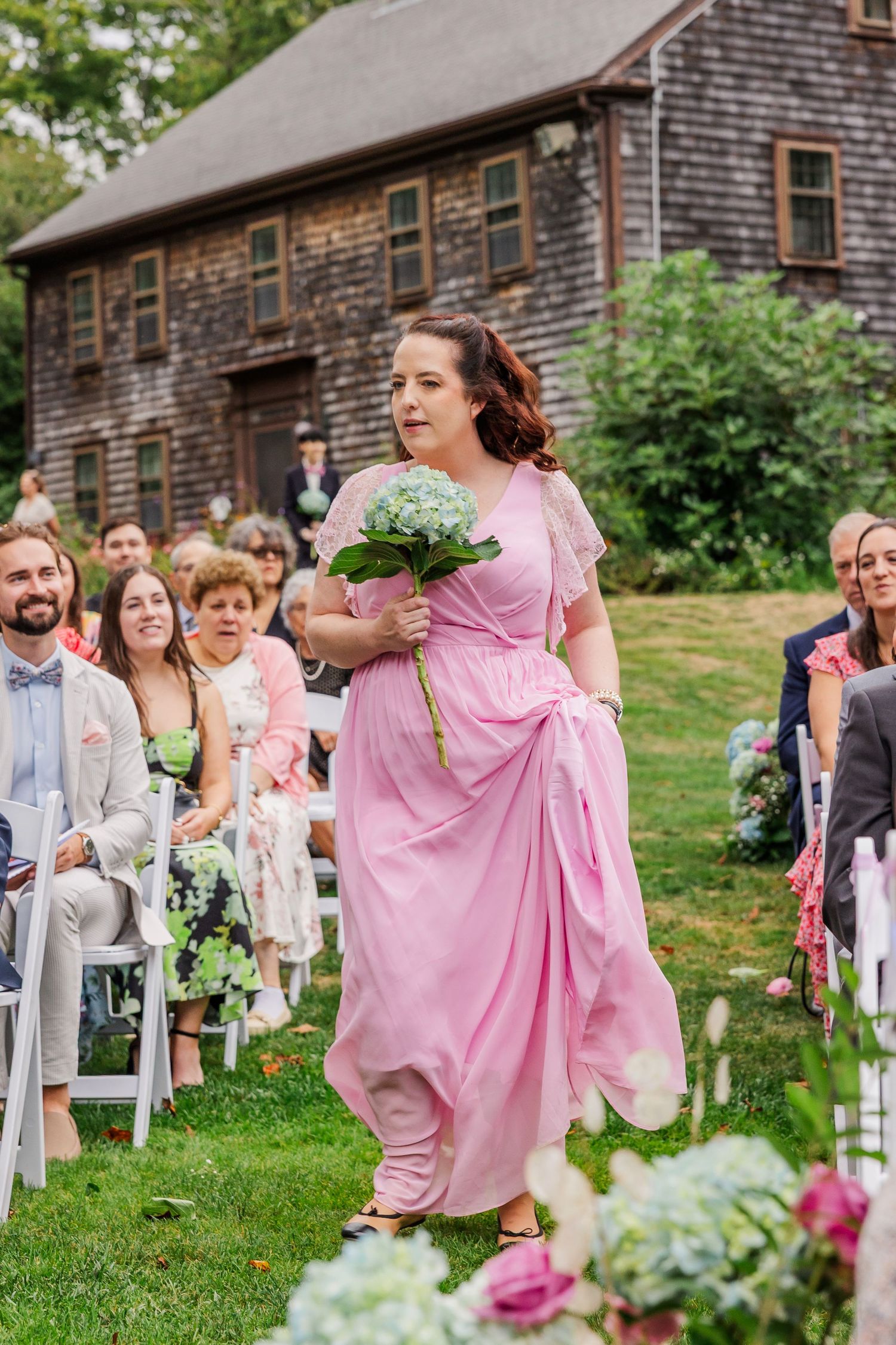 Wedding guests in pink dresses walk down an outdoor aisle lined with white chairs at a rustic barn ceremony.