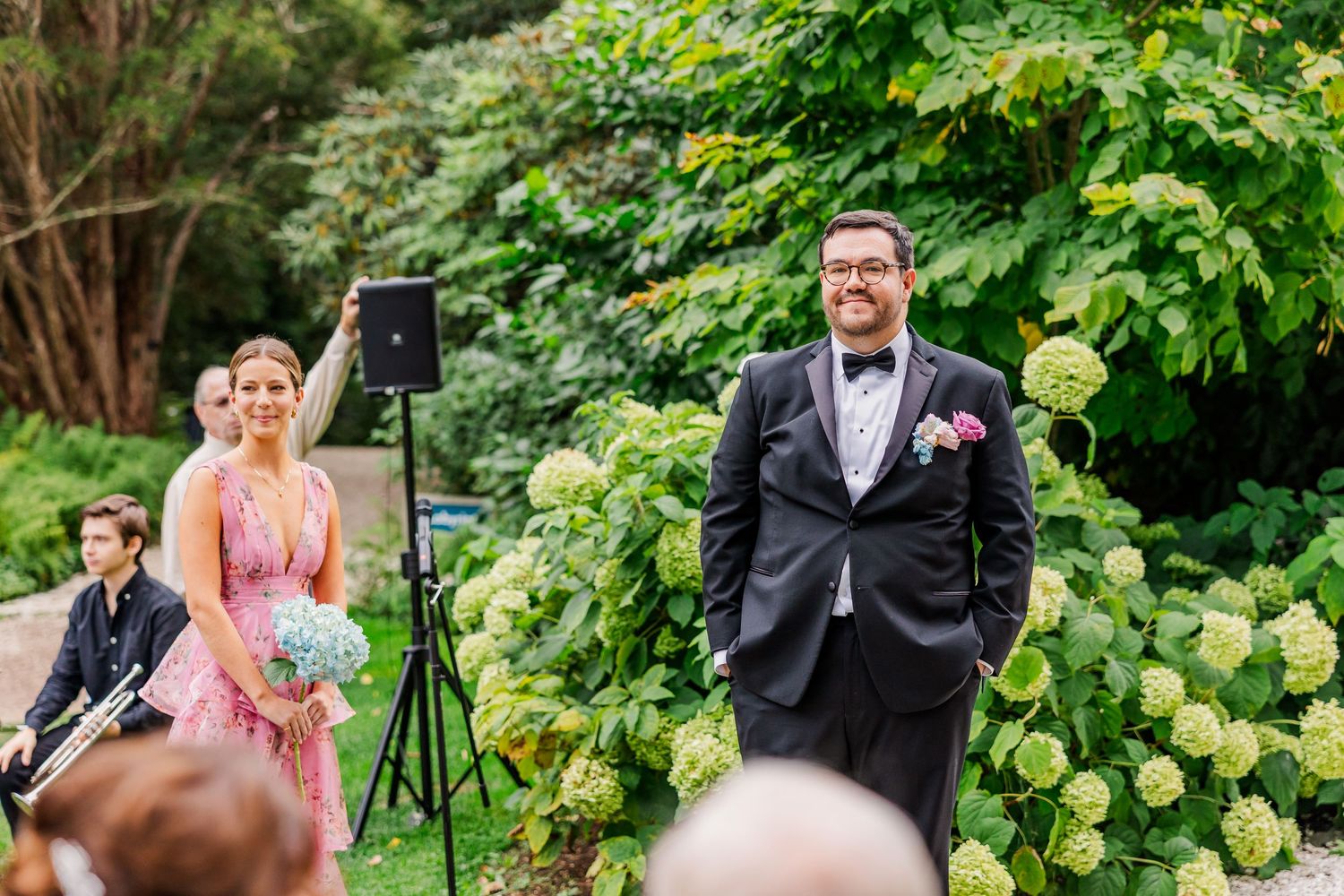 Outdoor wedding ceremony in a lush garden setting with hydrangea bushes and professional photography equipment.