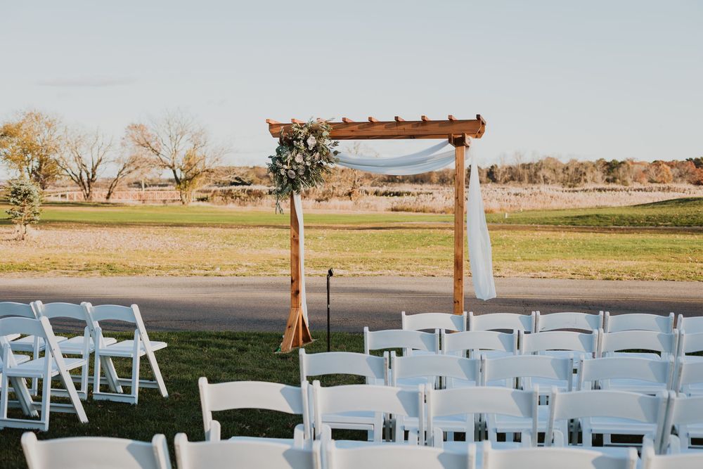 White chairs arranged for an outdoor wedding ceremony with a rustic wooden arch and scenic countryside backdrop.