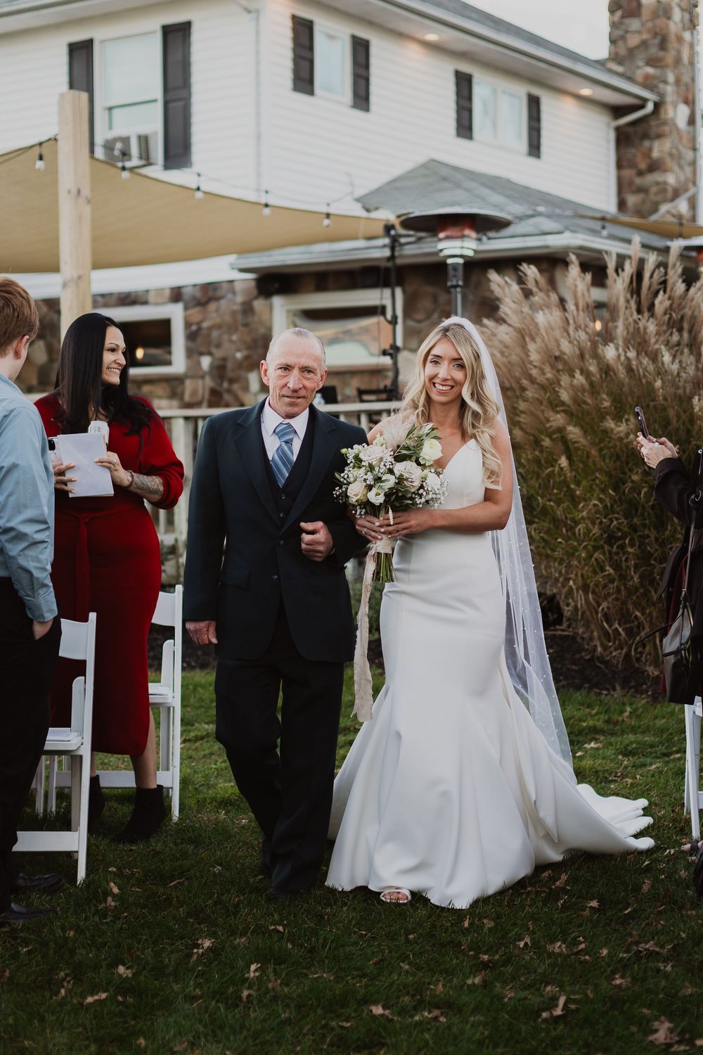 A wedding ceremony outdoors with guests and decorative ornamental grasses visible in the background.