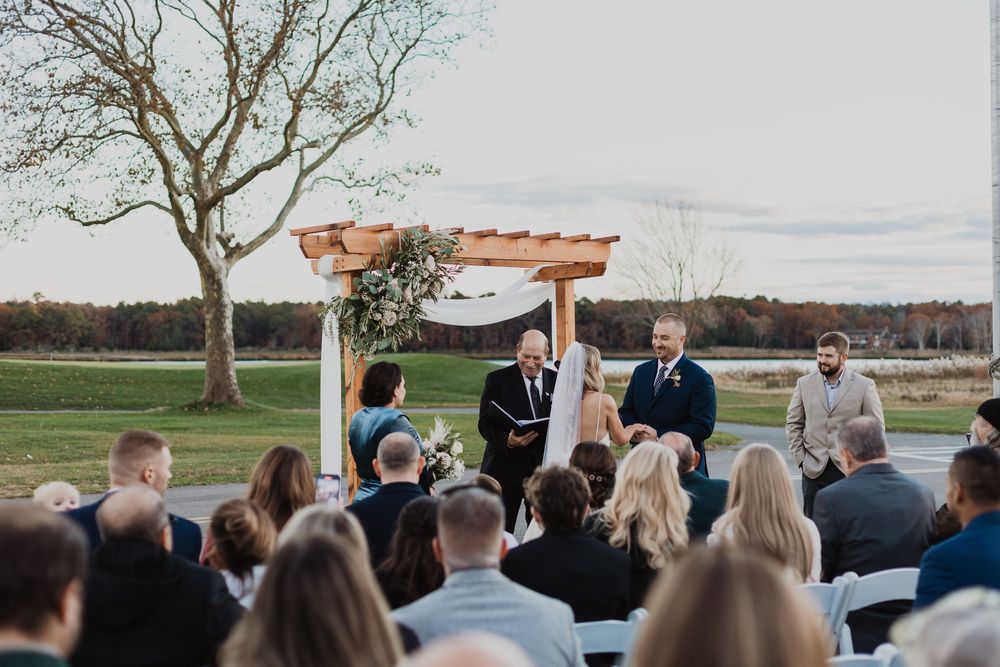 Outdoor wedding ceremony in progress with guests seated on white chairs facing a wooden pergola against a natural landscape.