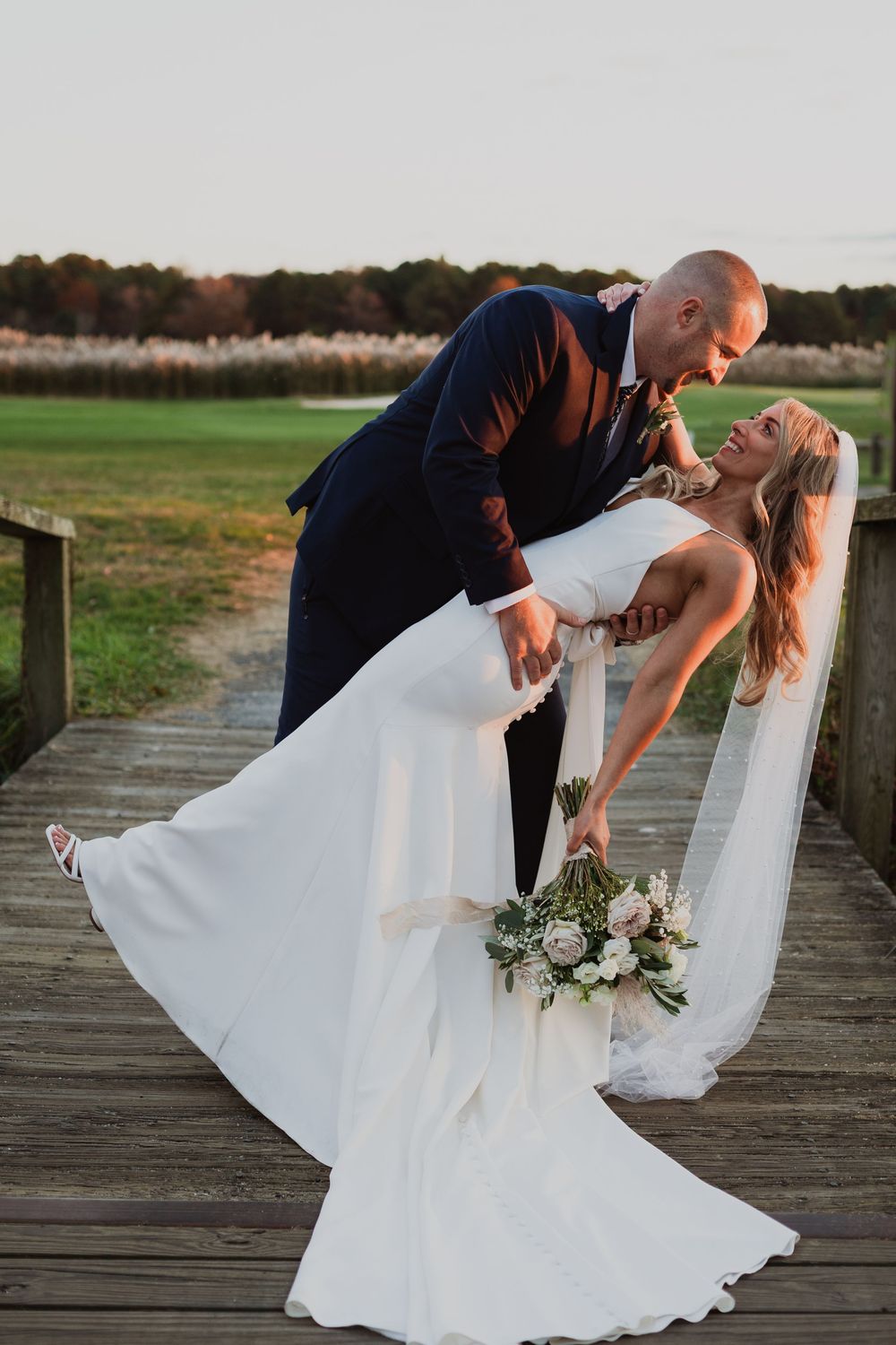 A couple in wedding attire share a romantic dip pose on a wooden boardwalk during sunset.