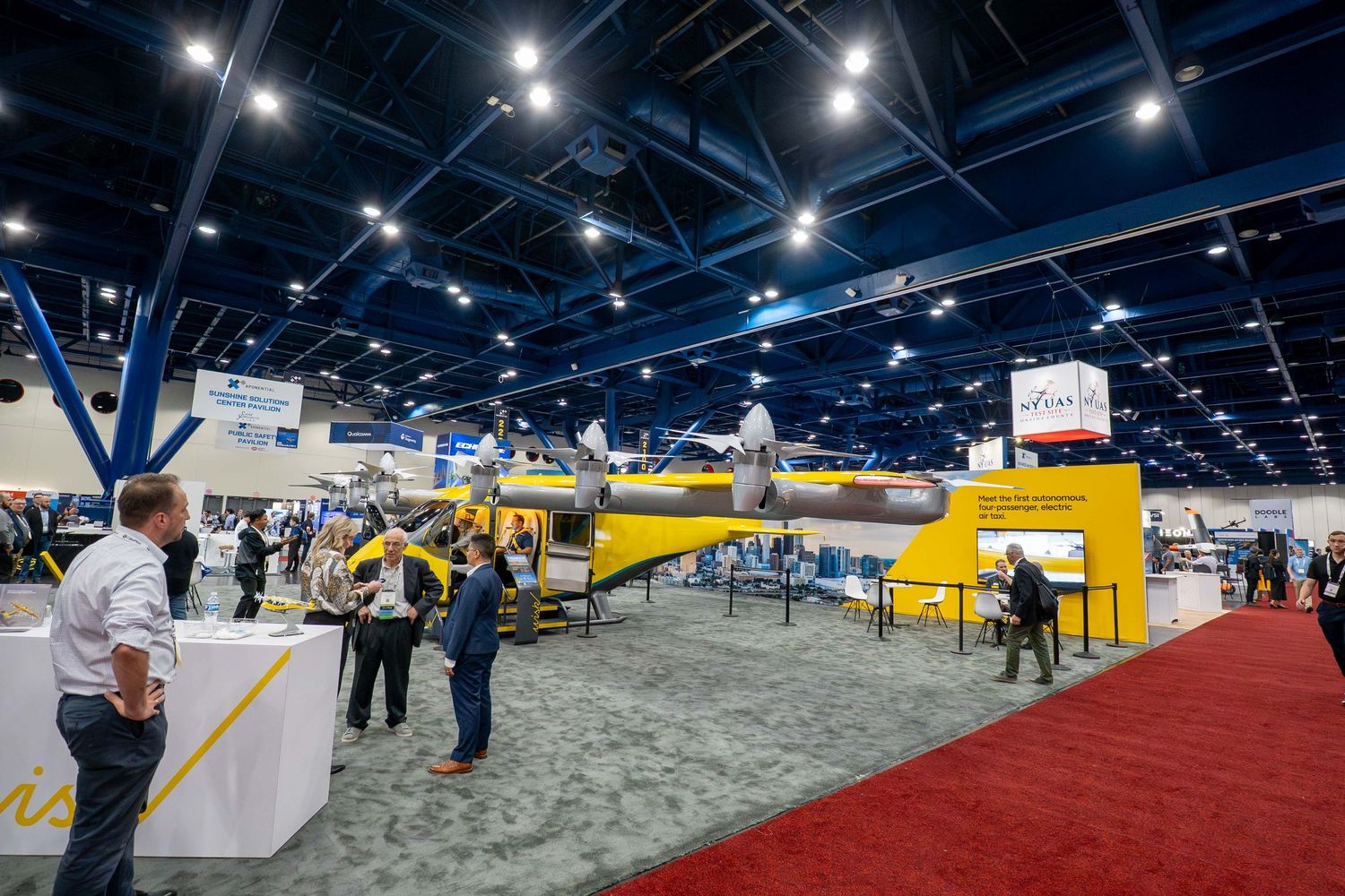 Large indoor convention center space with exhibitor booths and displays set up across a carpeted floor under bright lighting.
