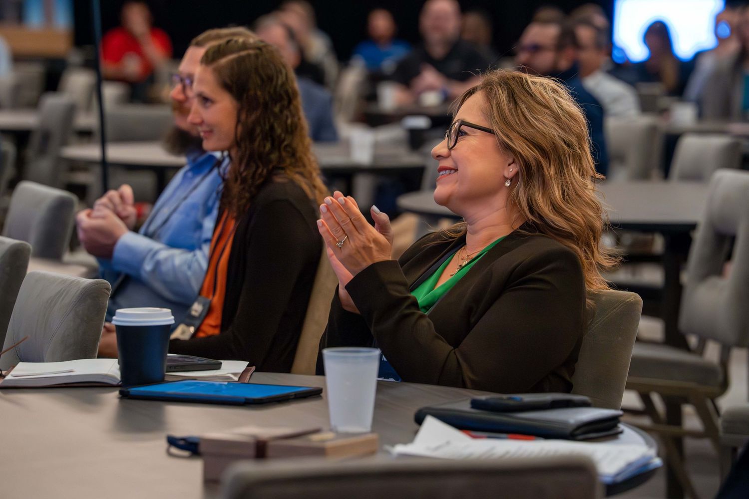 Audience members sit in rows of chairs at a conference or meeting, taking notes and listening attentively.